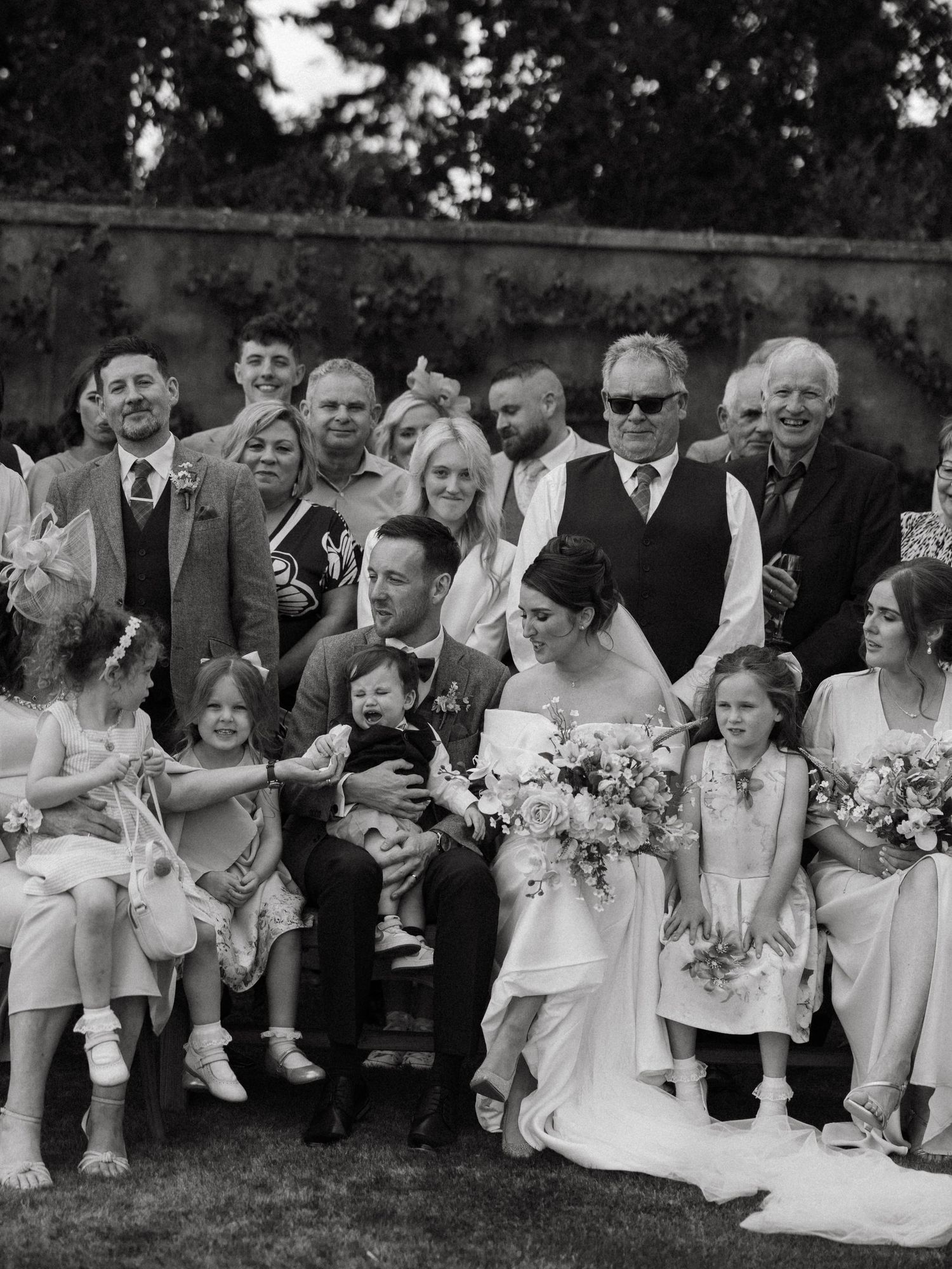 Wedding party members seated on a bench outdoors share candid moments and laughter during a photo session.