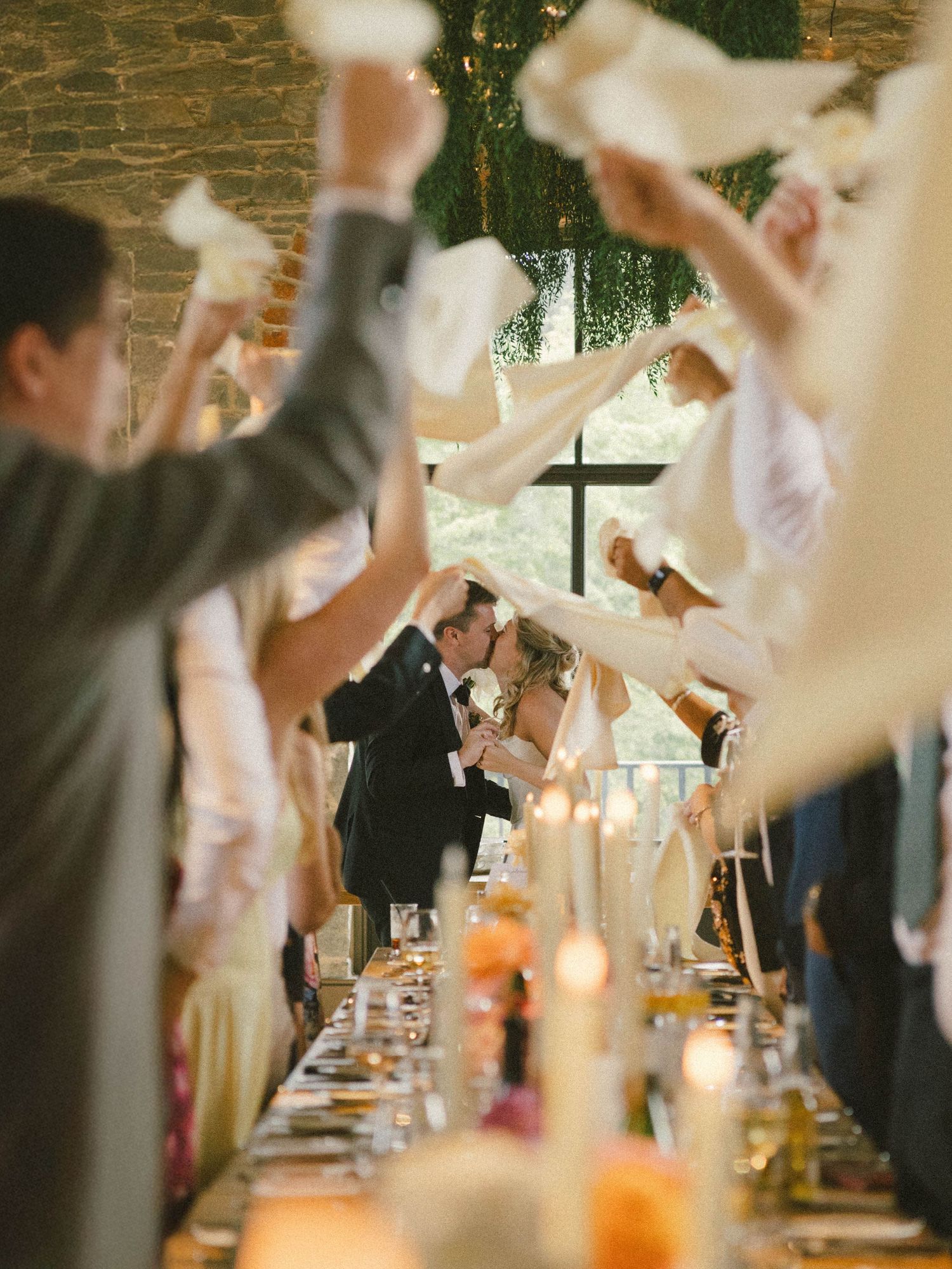 Wedding guests raise their glasses in celebration during a toast at an elegant reception with a cross visible in background.