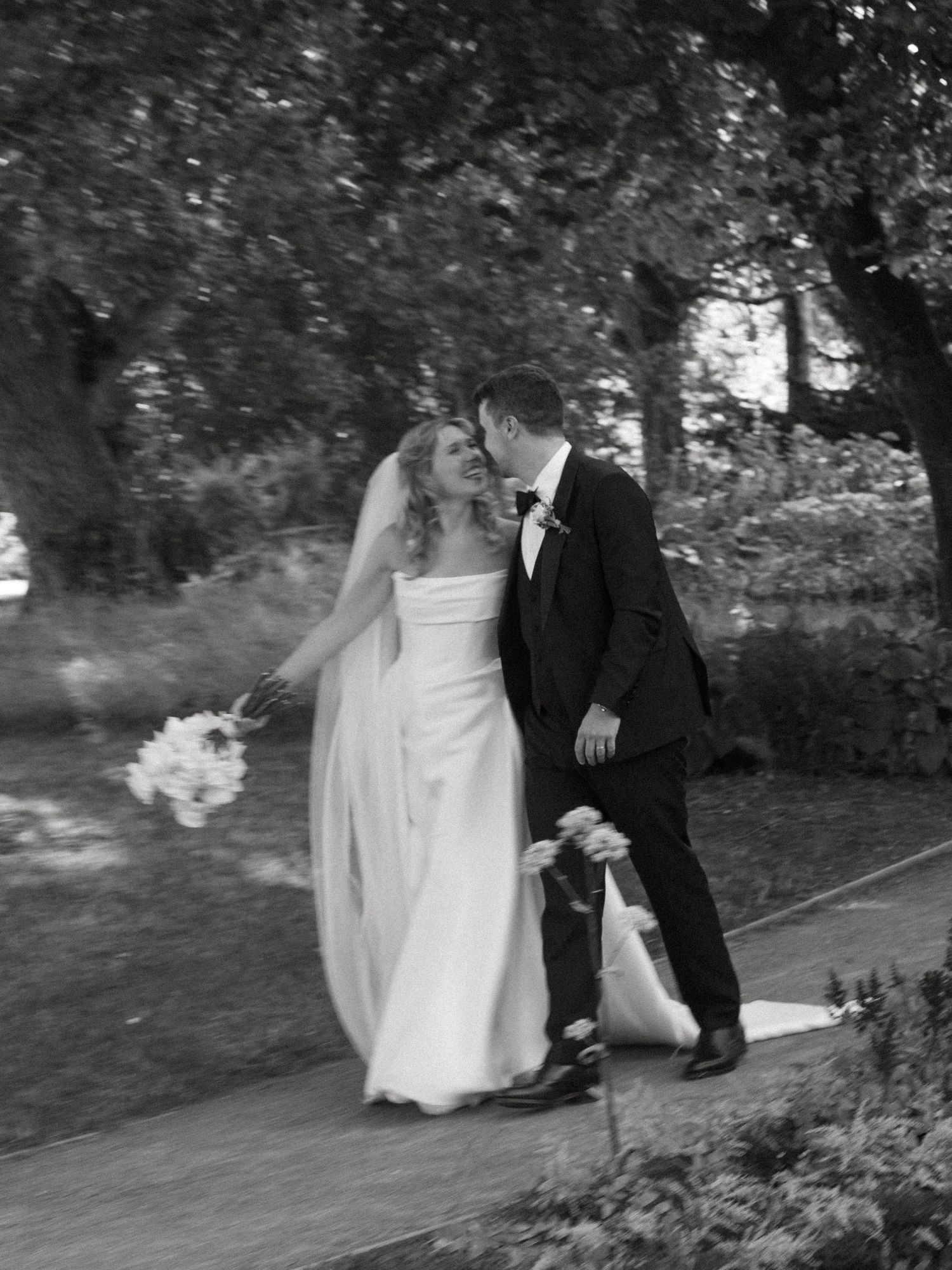 Black and white romantic wedding portrait of a couple walking along a garden path.