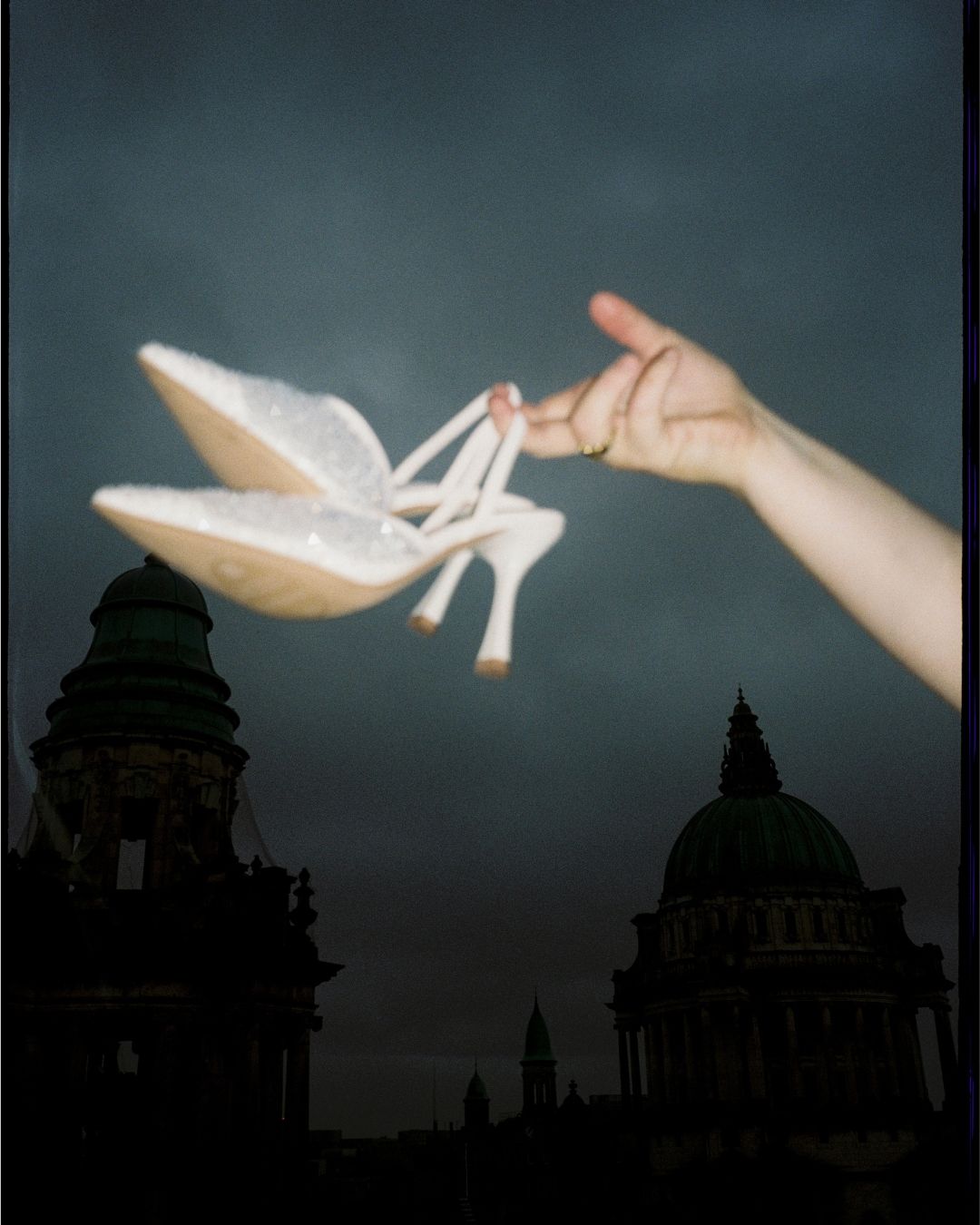 A white high heel shoe floats mysteriously against a dark background with architectural silhouettes visible.