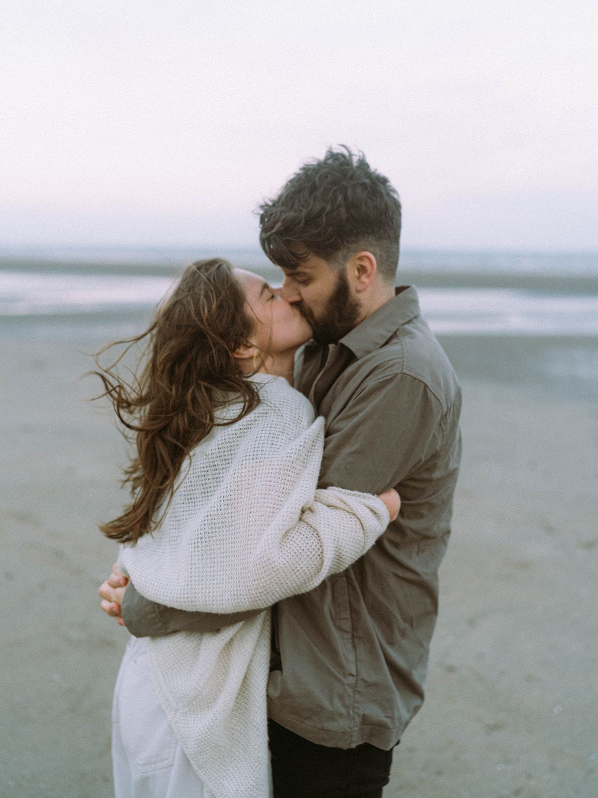 Couple in cozy sweaters sharing tender kisses on a misty beach.