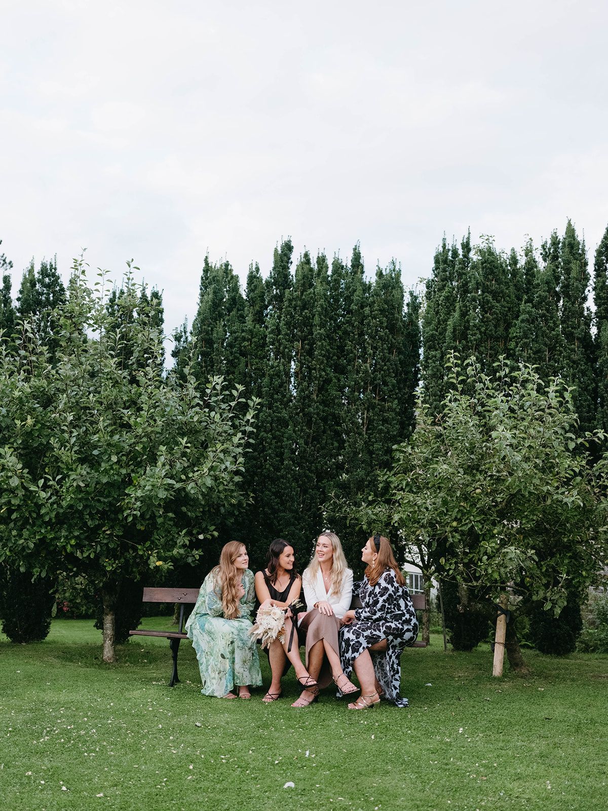 A group sits together on a bench surrounded by tall evergreen trees and lush greenery in a peaceful garden setting.