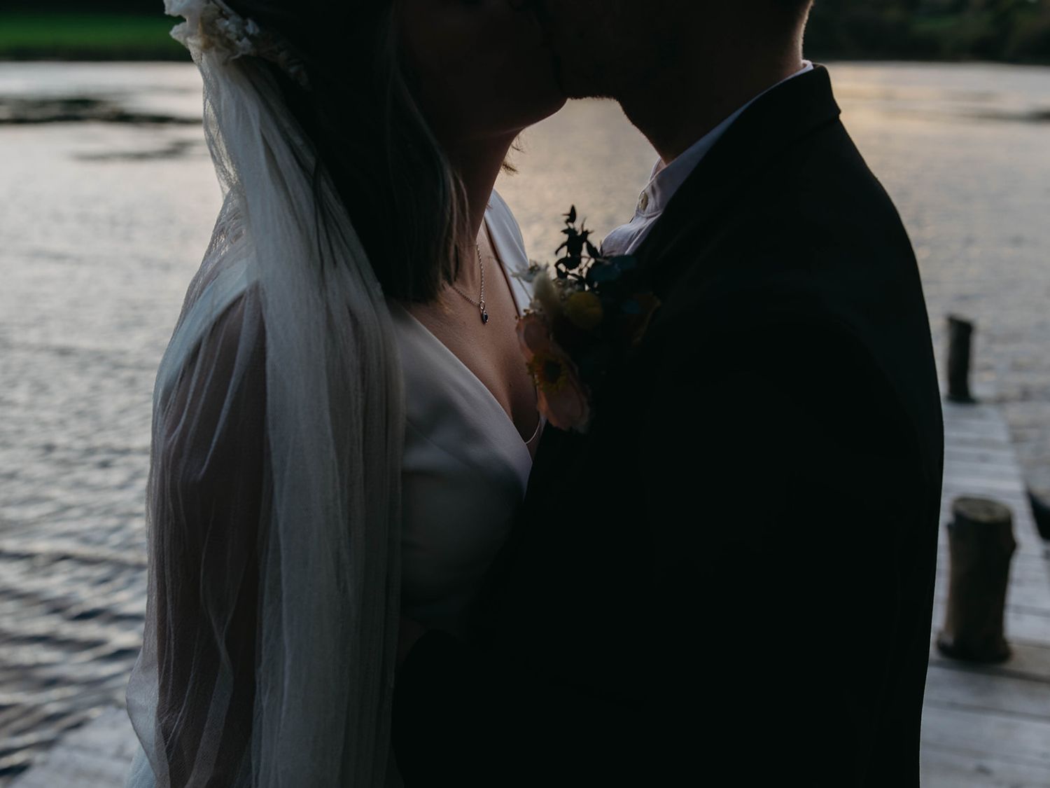 A close-up wedding photo silhouette against a waterfront at dusk showing a bride's veil and groom's suit.