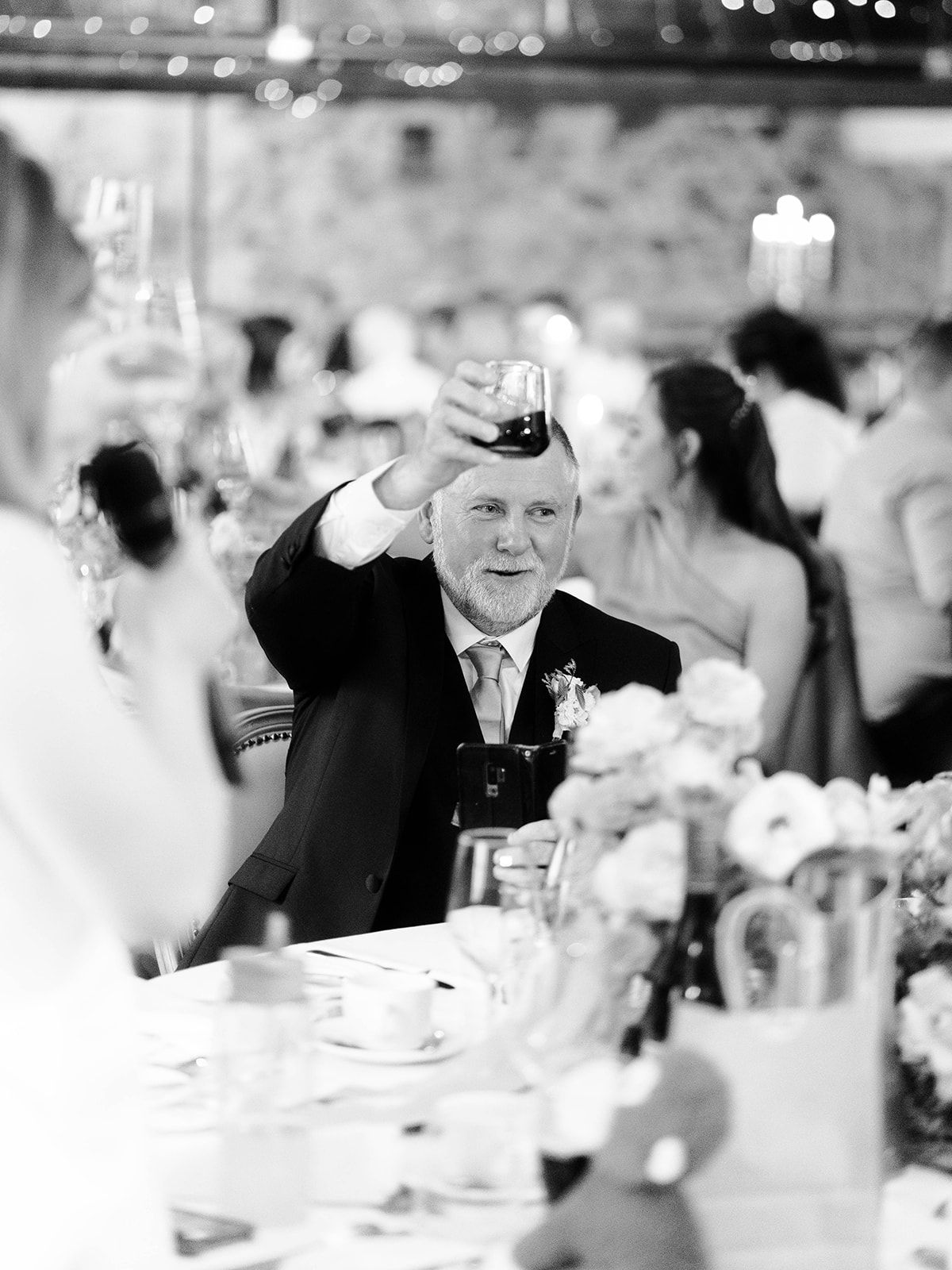 A wedding guest raises their glass in a toast at an elegant reception with bokeh lights and white decor in the background.