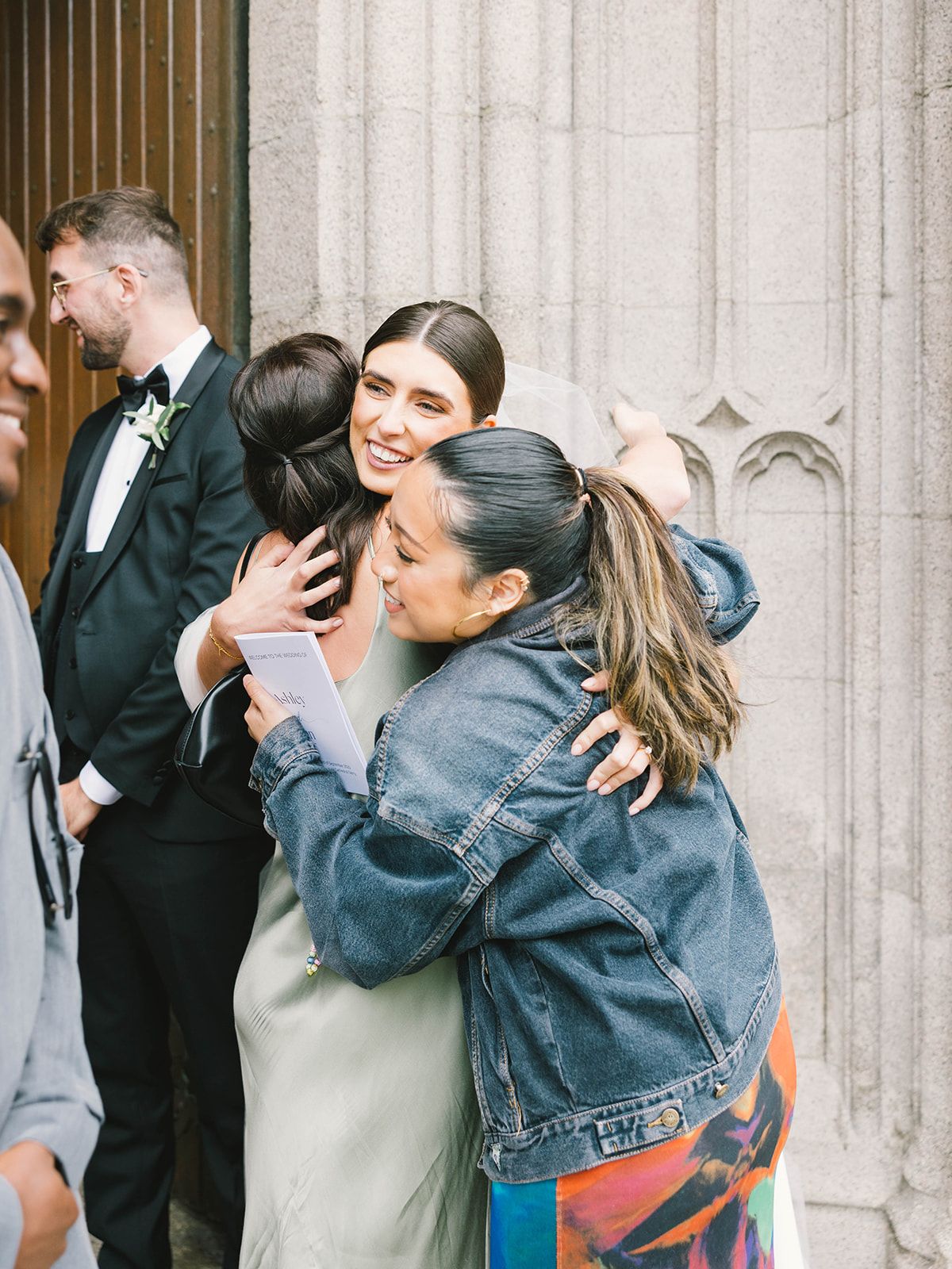Two people share a warm embrace and laugh together outside a building with stone columns in formal attire.