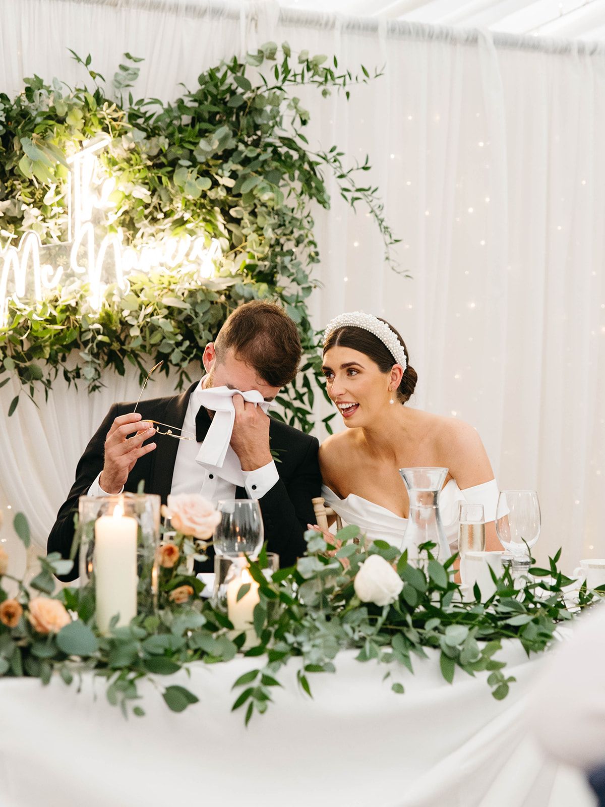 A couple sharing a joyful moment at their wedding reception table decorated with lush greenery and glowing candles.