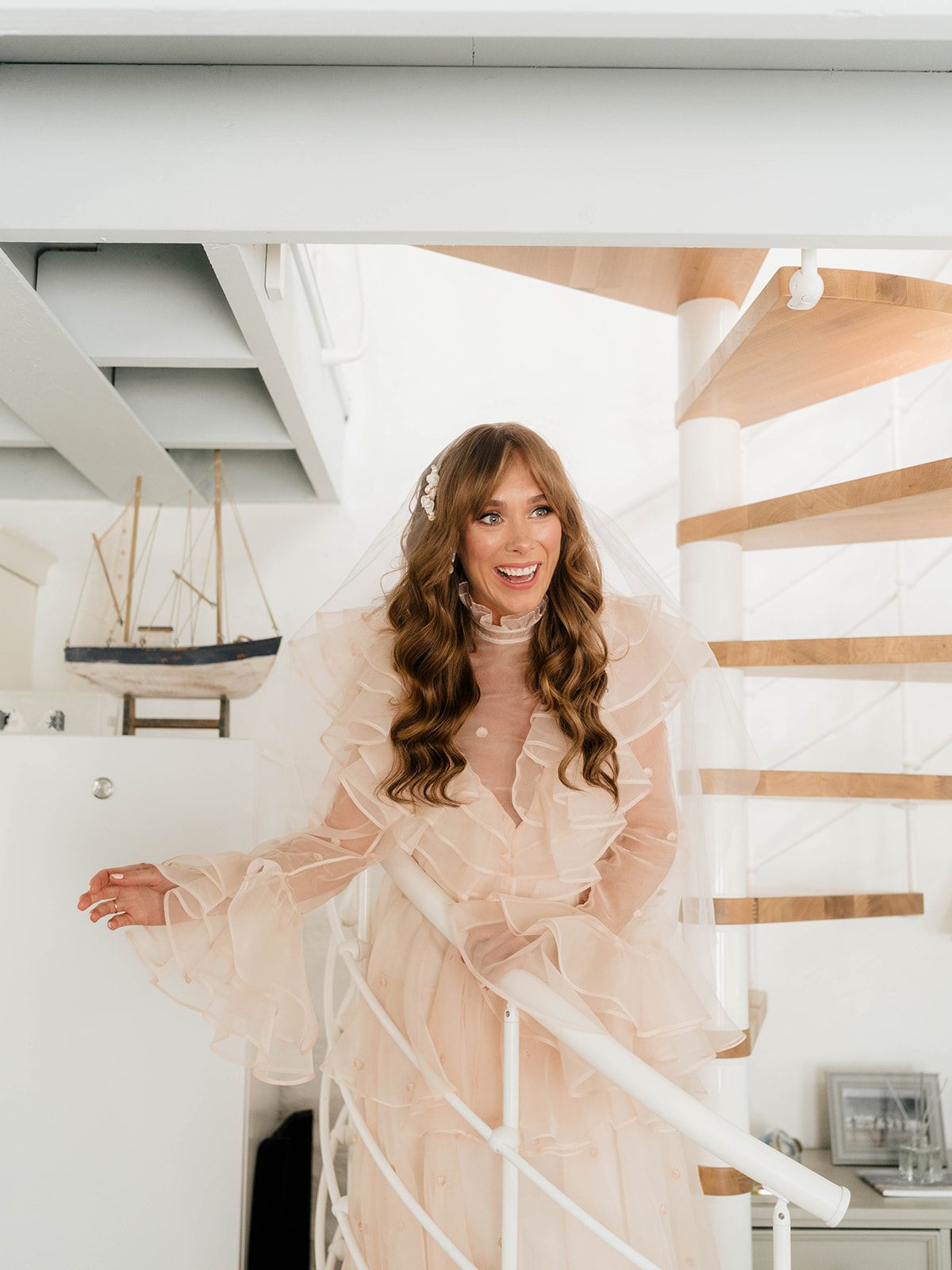 Woman in flowing peach dress poses elegantly on white spiral staircase with nautical decor in background.