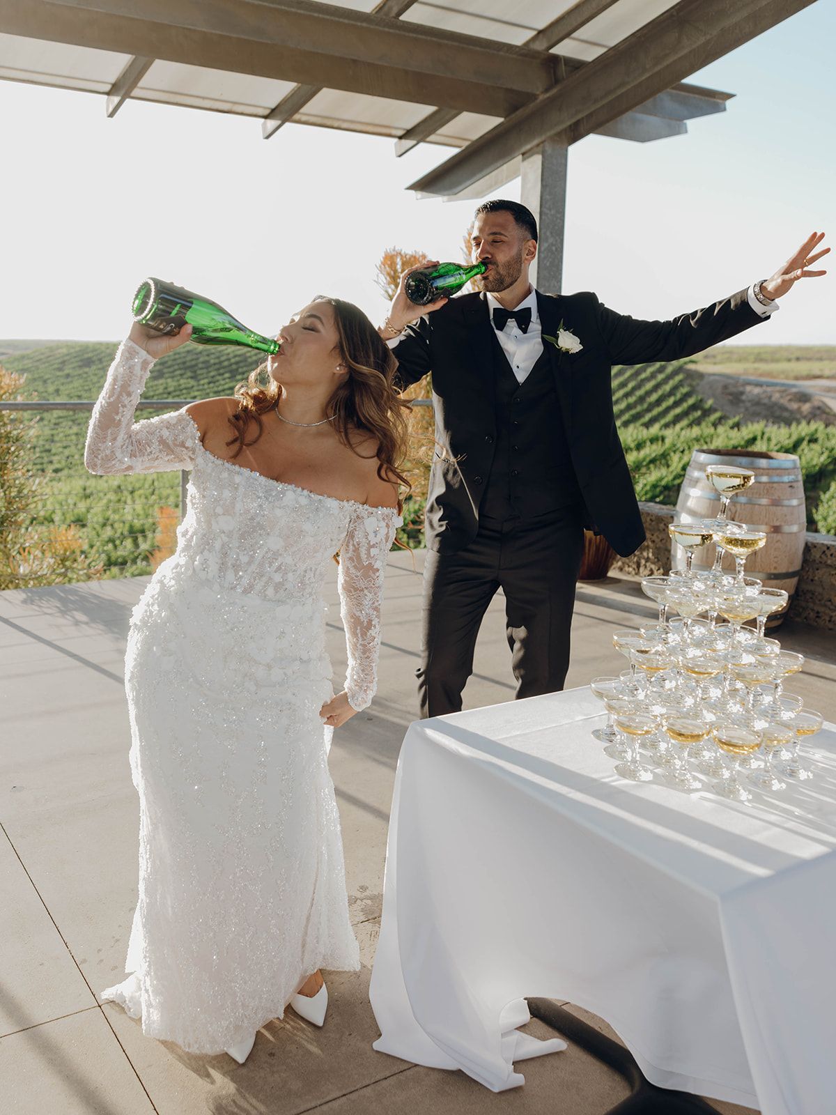 A couple celebrates their wedding toast next to a champagne tower at a scenic vineyard venue.