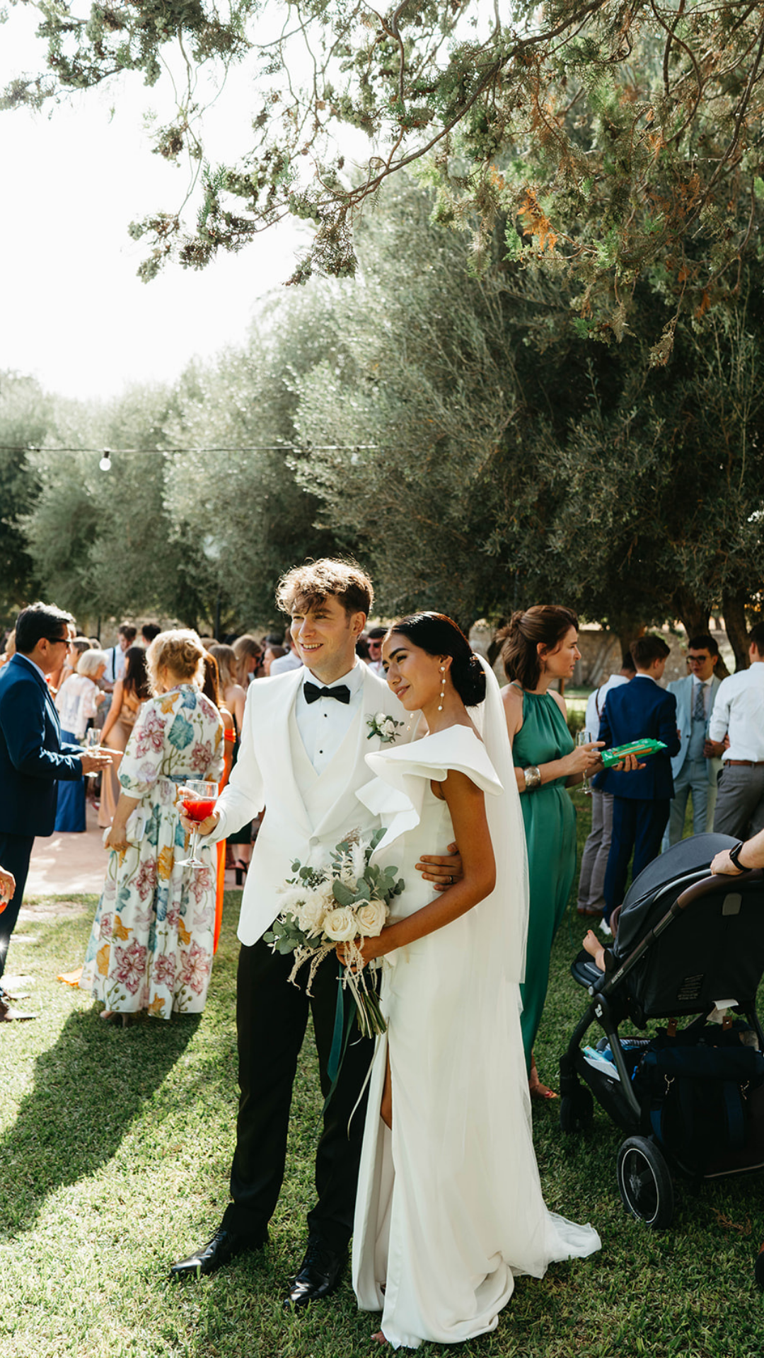 Sunny outdoor wedding reception with guests mingling under trees.
