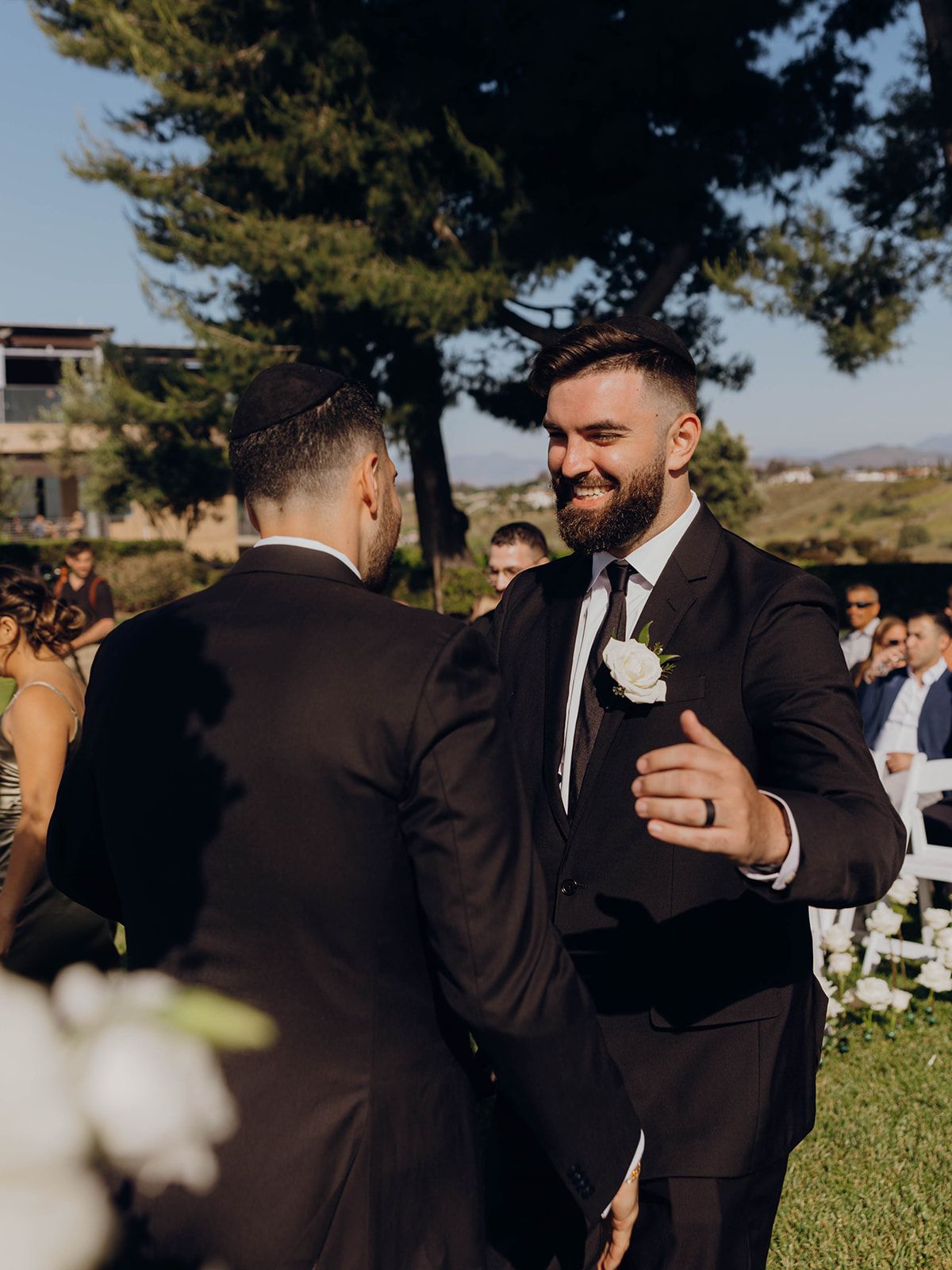 Two grooms in matching dark suits share a joyful moment during an outdoor wedding ceremony.