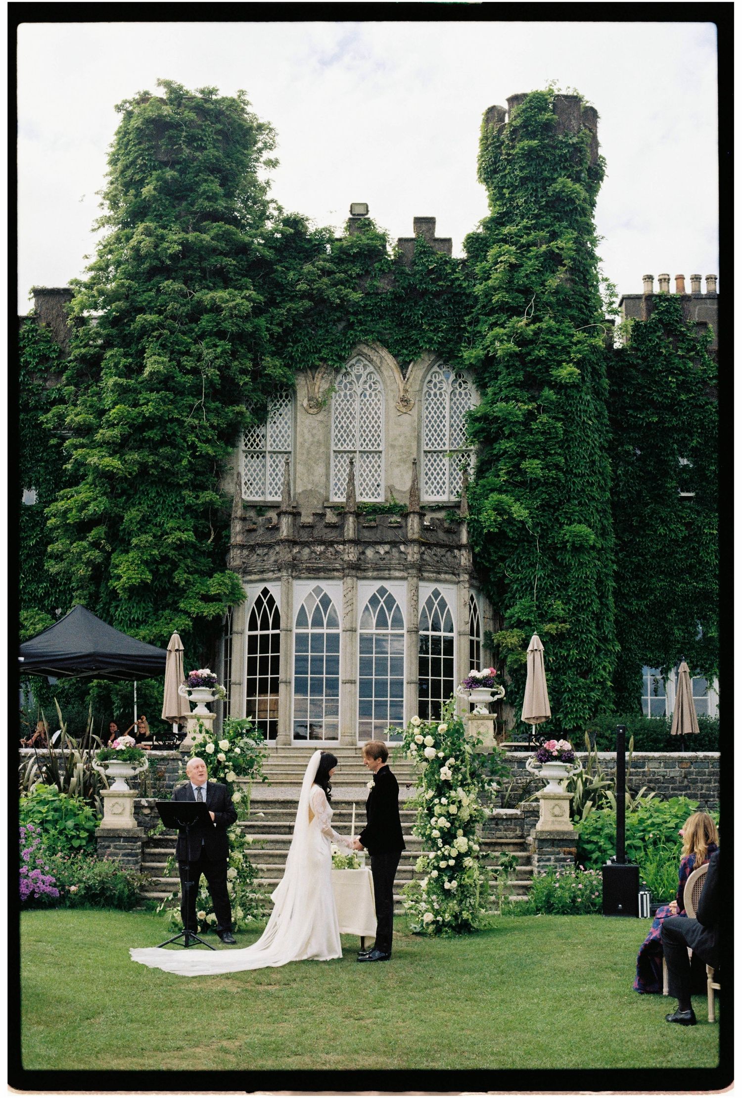 Outdoor wedding ceremony in front of an ivy-covered Gothic castle with floral arch and bridal party on lush green lawn.