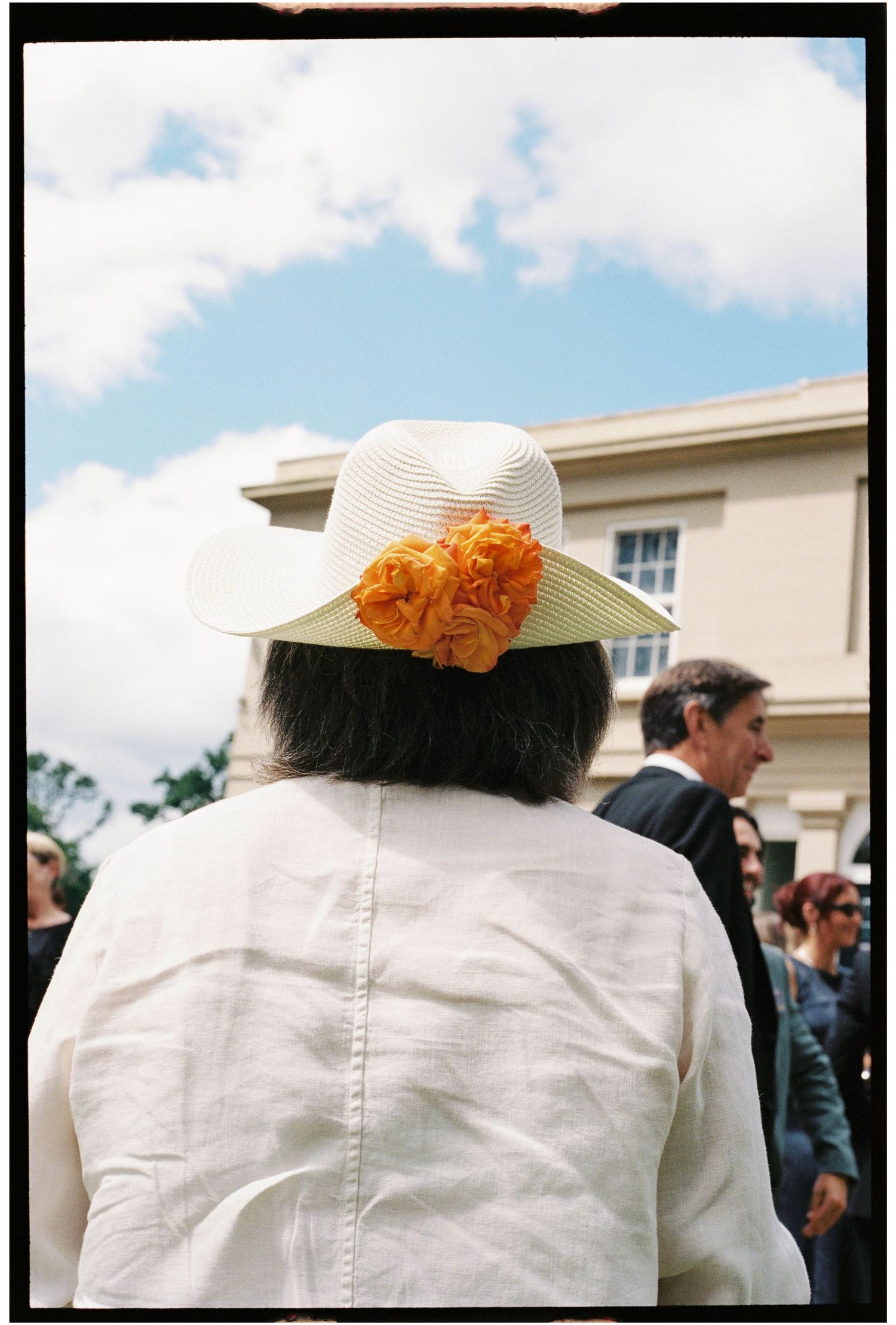 Woman wearing white hat with orange floral decoration at an outdoor wedding celebration.