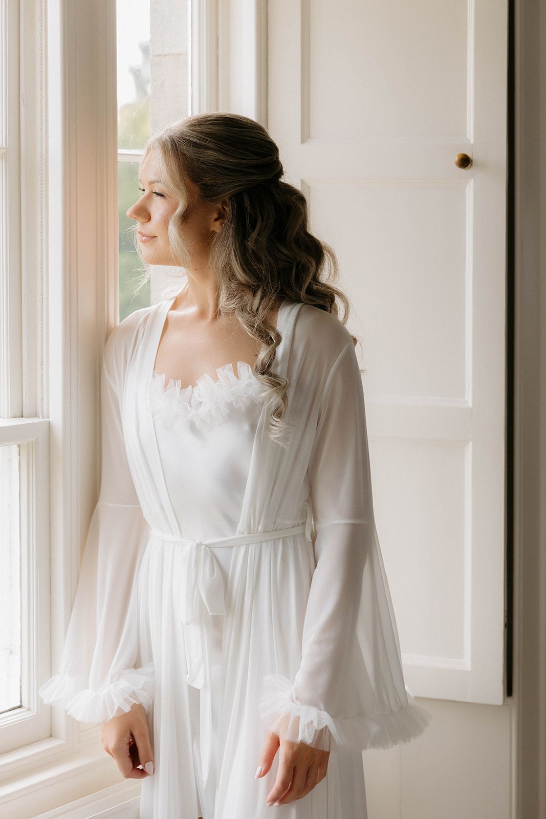 Woman in flowing white dress and robe stands near bright window in elegant boudoir setting.