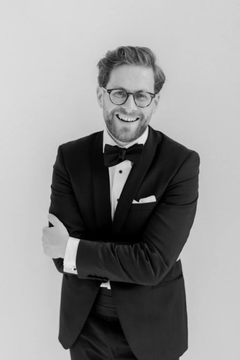 Smiling man in black tuxedo and bow tie with glasses, arms crossed, black and white portrait photo.