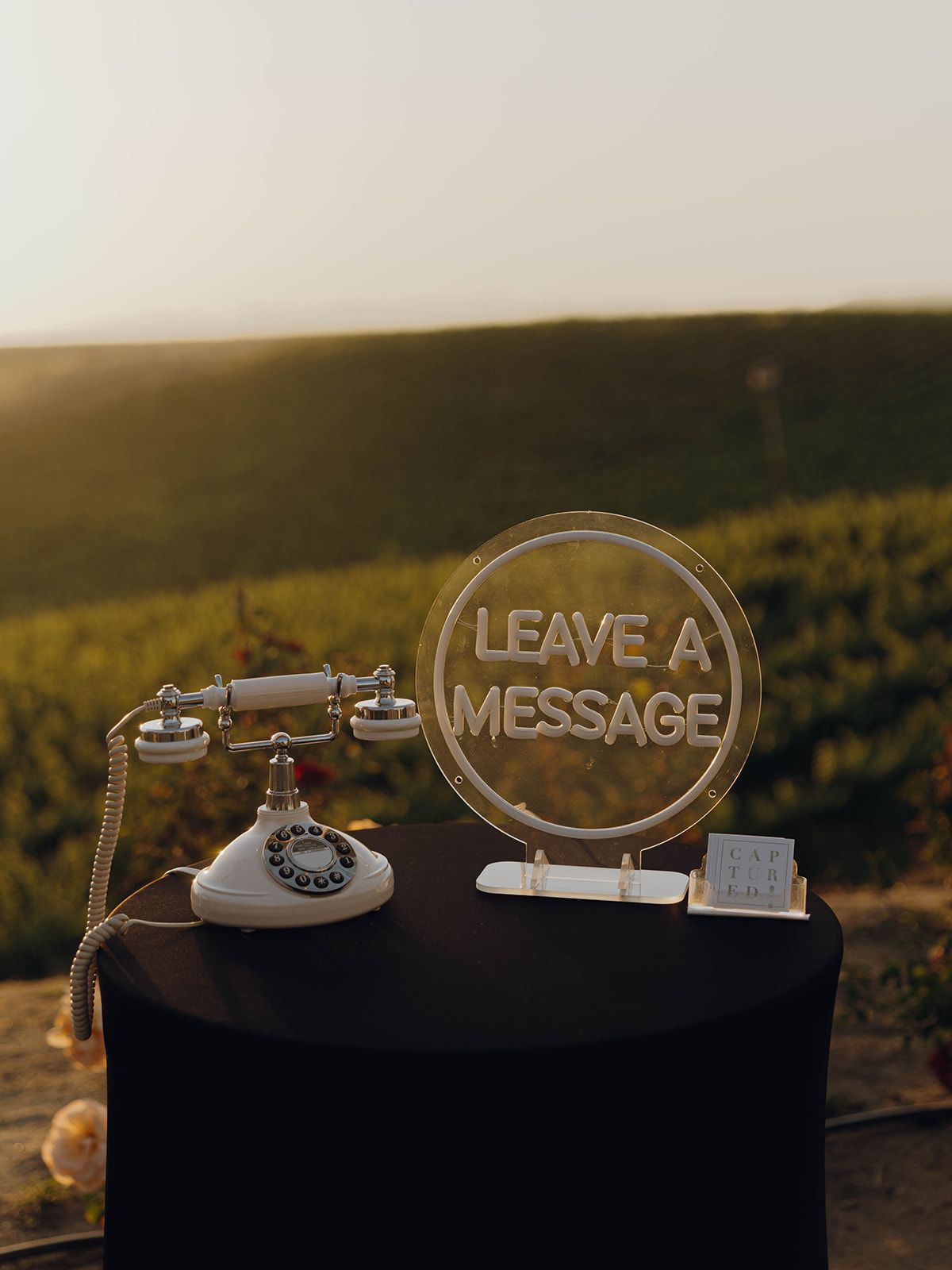 Vintage rotary phone and 'Leave a Message' sign displayed on a black surface against a sunset landscape backdrop.