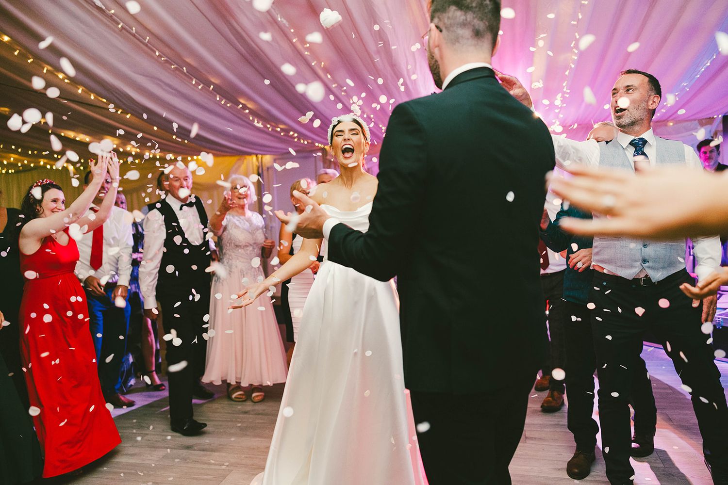 Festive wedding reception with dancing couples surrounded by floating bubbles under pink-lit draped fabric ceiling.