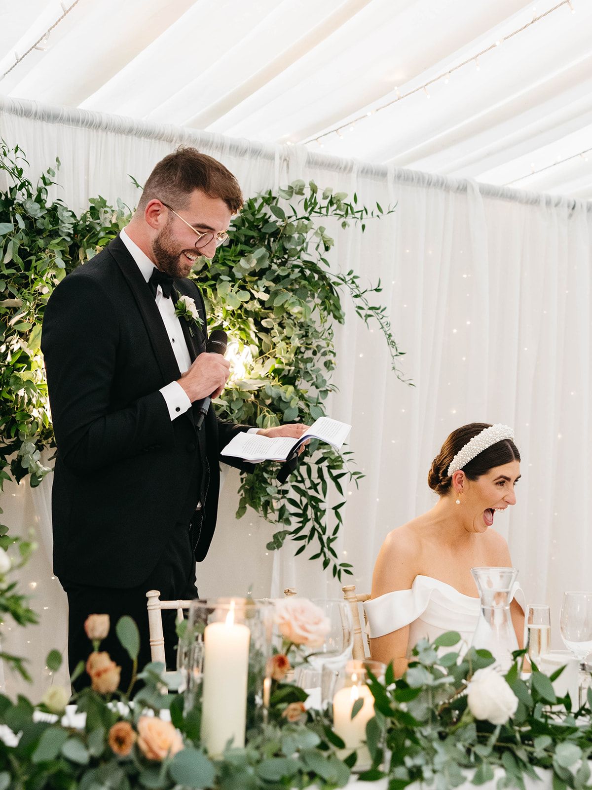 A wedding reception under a white tent with floral greenery arrangements, tall candles and elegant table settings.