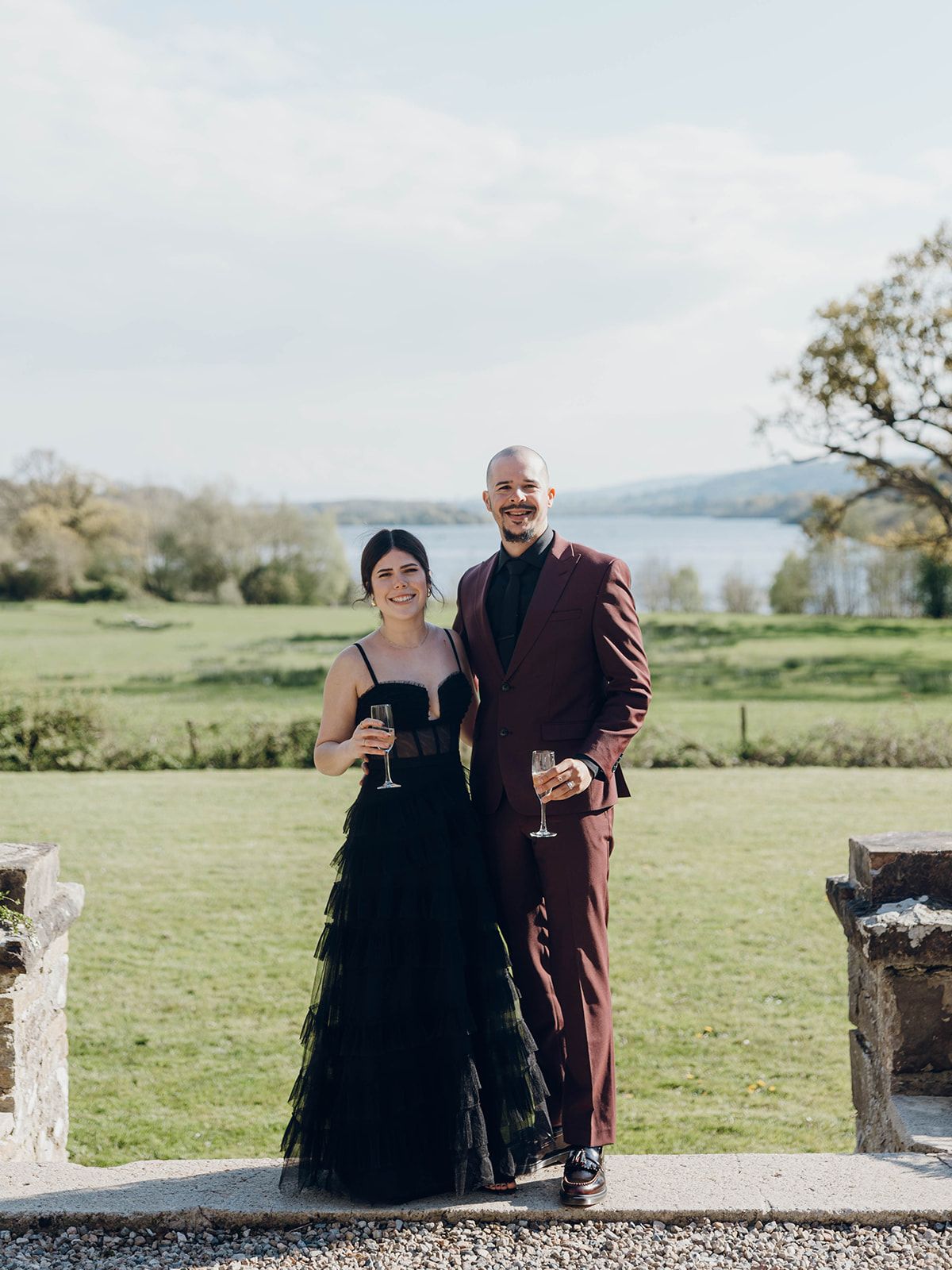 Couple in formal attire stands on grassy hillside grounds with scenic lake view, holding wine glasses at outdoor event.