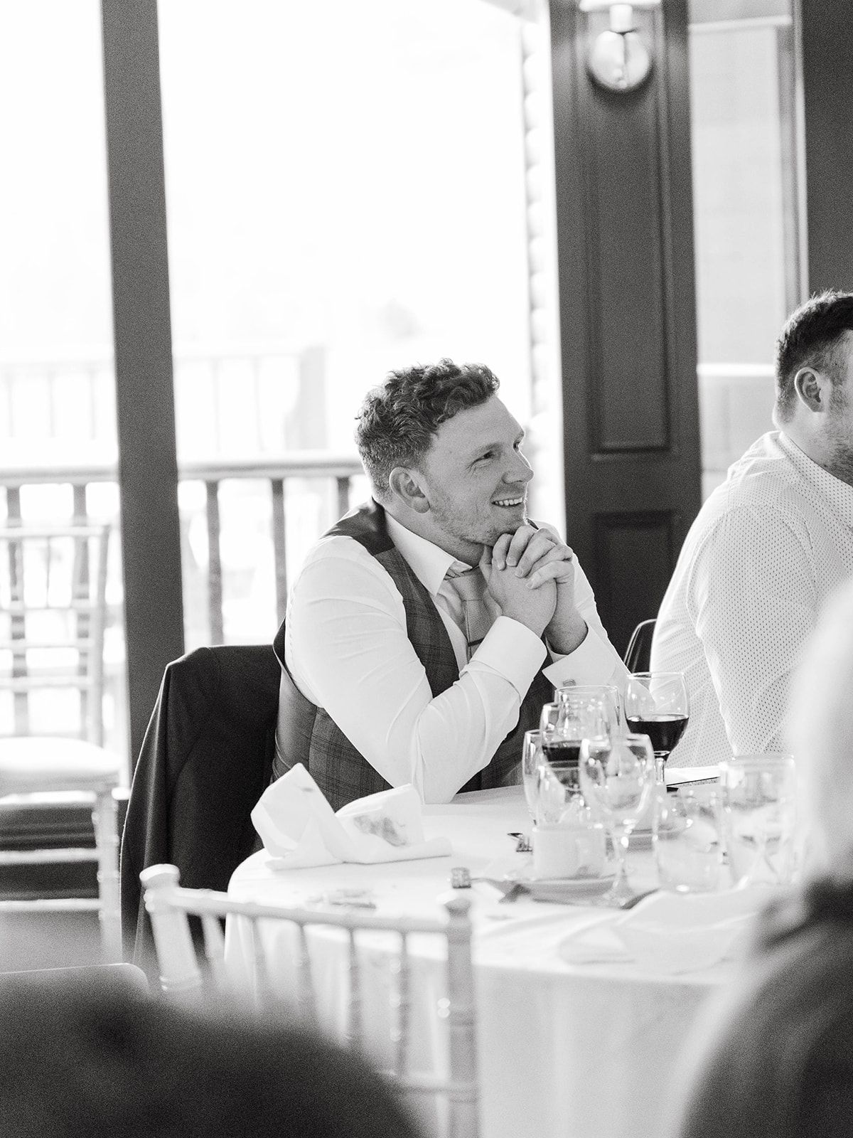 A man in formal attire smiles while sitting at an elegant table setting with wine glasses and white linens.