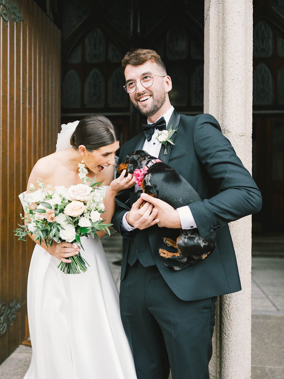 A joyful wedding couple laughs while holding their black dog during their wedding portraits at a church.