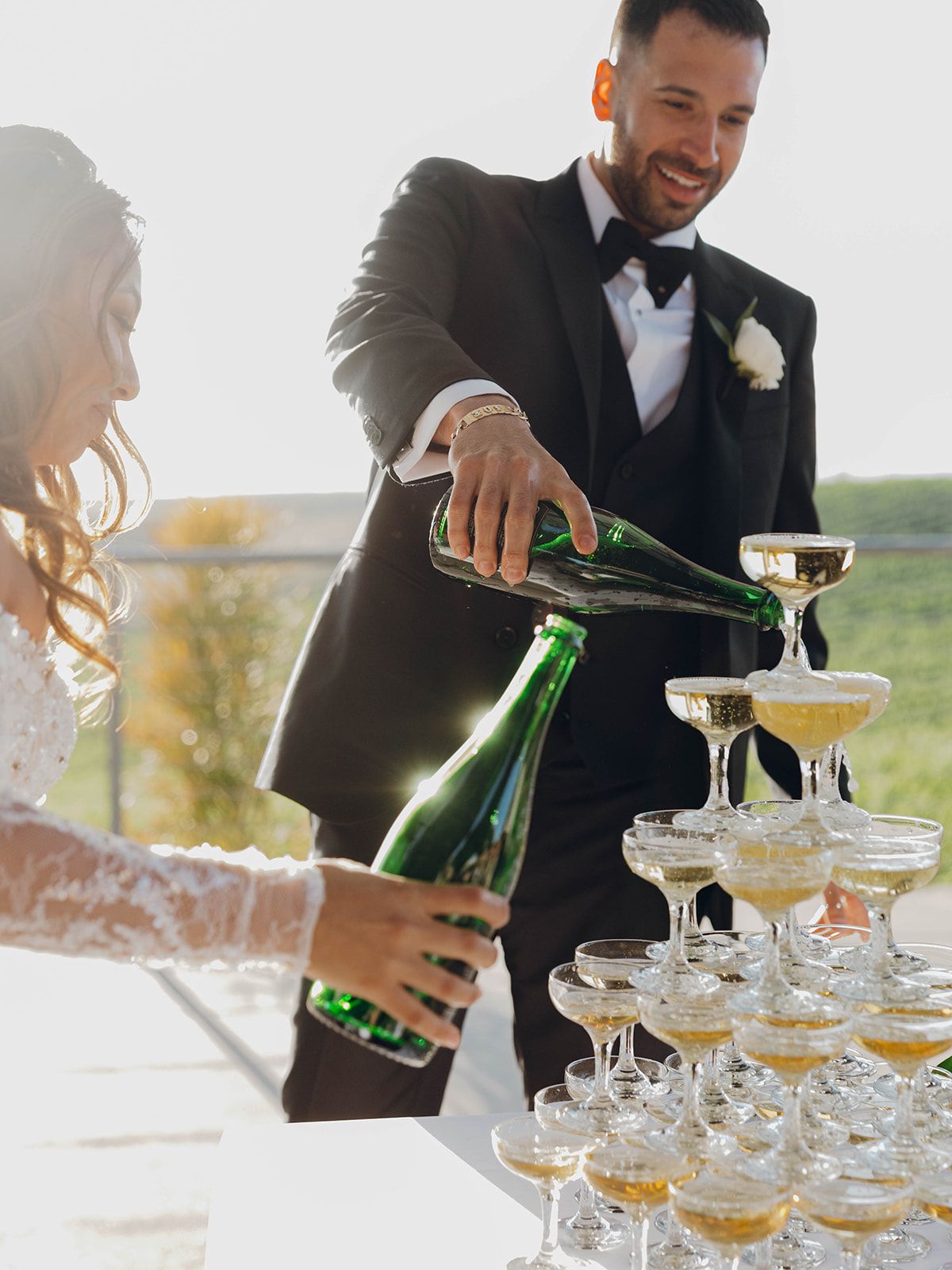 A person in a black tuxedo pours champagne into a tower of stacked coupe glasses at an elegant outdoor celebration.
