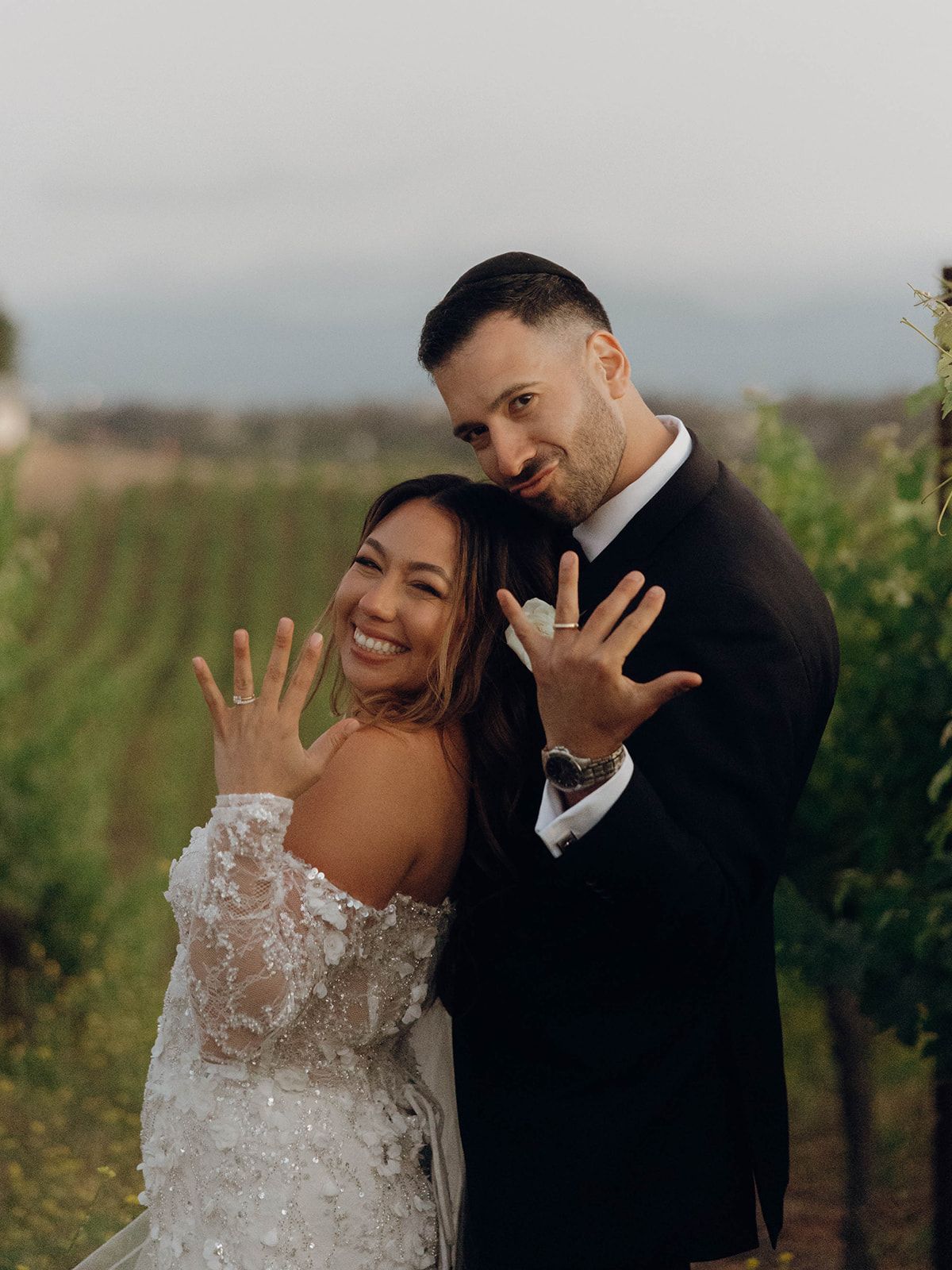 Happy newlyweds showing off their wedding rings together at a scenic vineyard in matching formal attire.