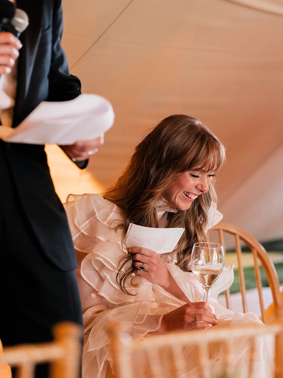 A bride in a ruffled dress laughs while seated at a wedding reception table.