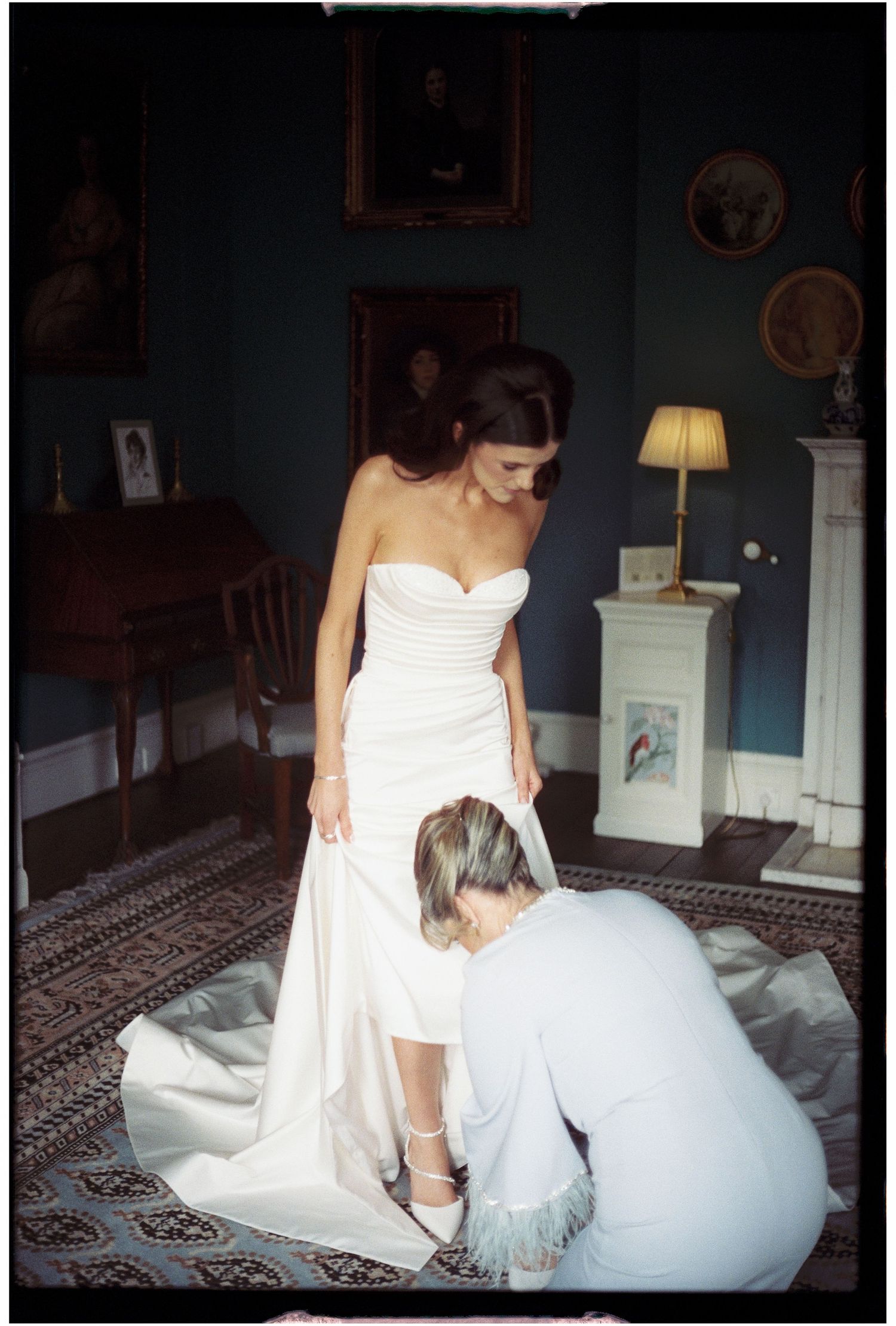A bride adjusts her white wedding gown while sitting in an elegantly furnished room.