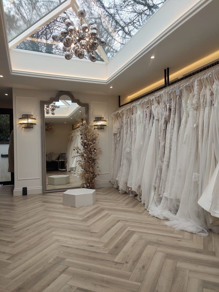 Elegant bridal boutique interior with herringbone floors, skylight, chandelier and rows of white wedding dresses on display.