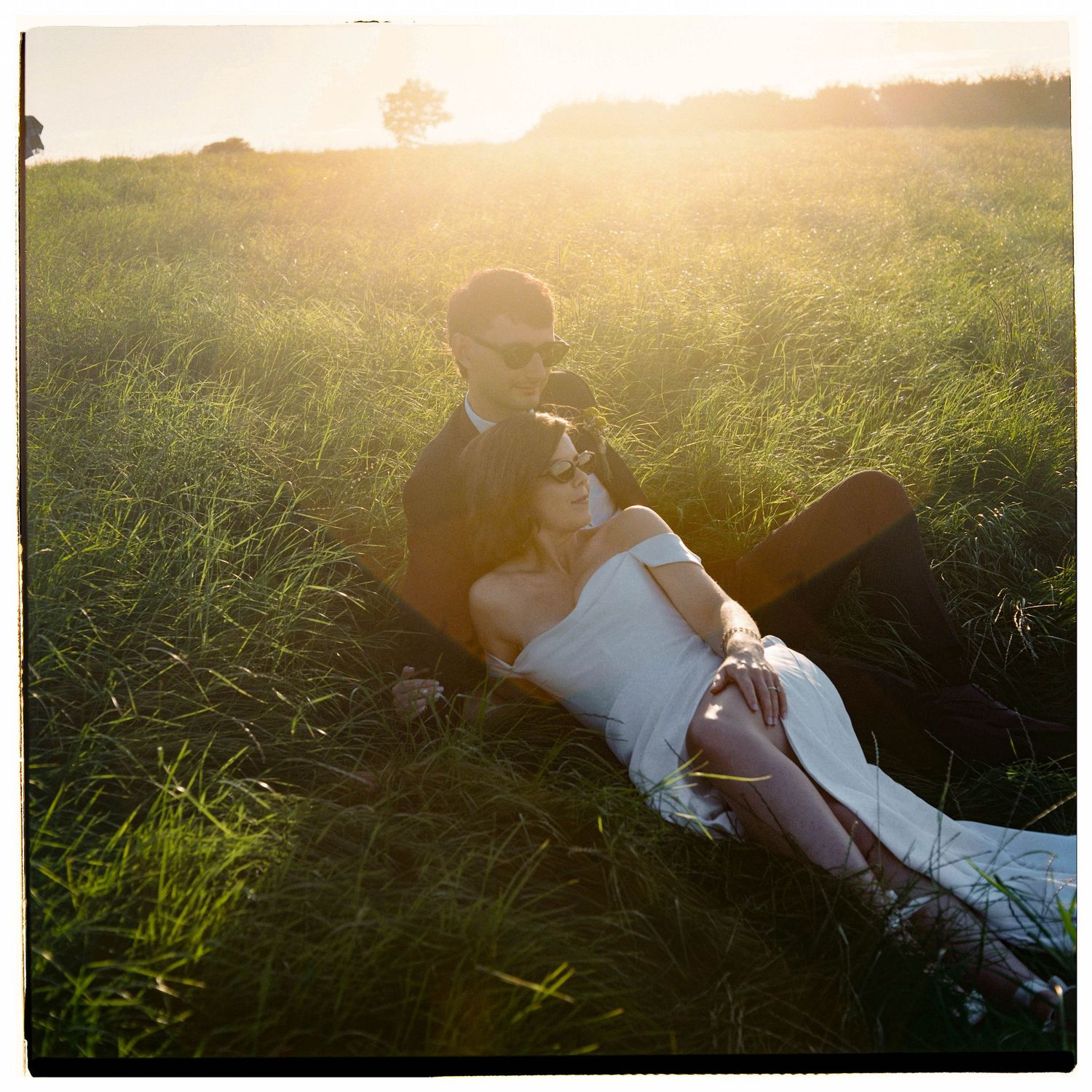 Couple relaxing in a sunlit green meadow at golden hour, woman in white dress leaning on man wearing sunglasses.