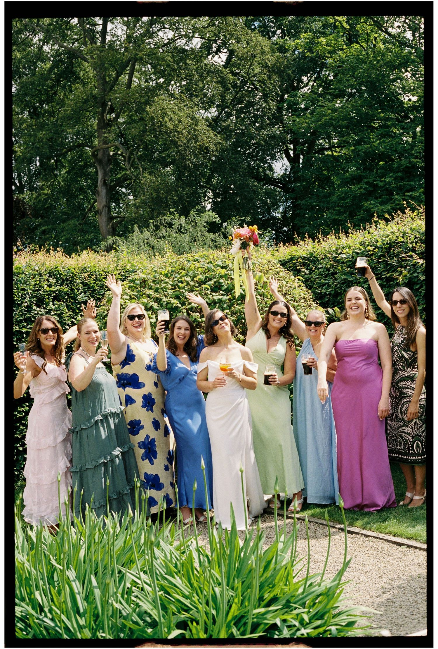 Bridesmaids in colorful mismatched gowns raise glasses in a lush garden setting during an outdoor wedding celebration.