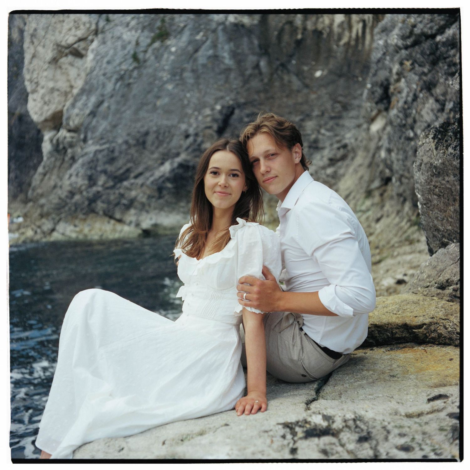 Couple in white attire sitting on rocky coastal cliffs, posing for a romantic engagement or wedding photo.