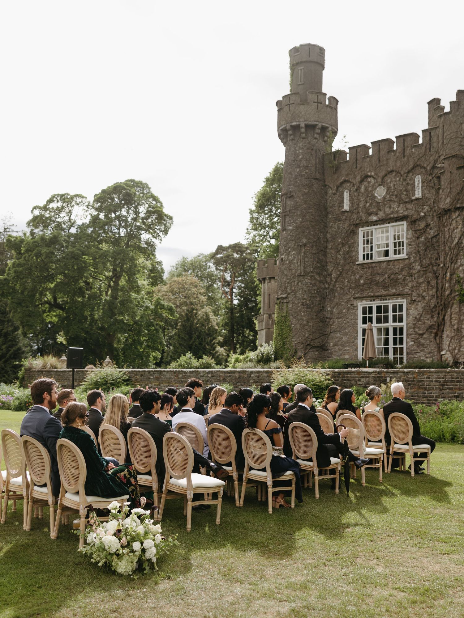 Outdoor wedding ceremony with guests seated on wooden chairs facing a historic stone castle surrounded by lush greenery.