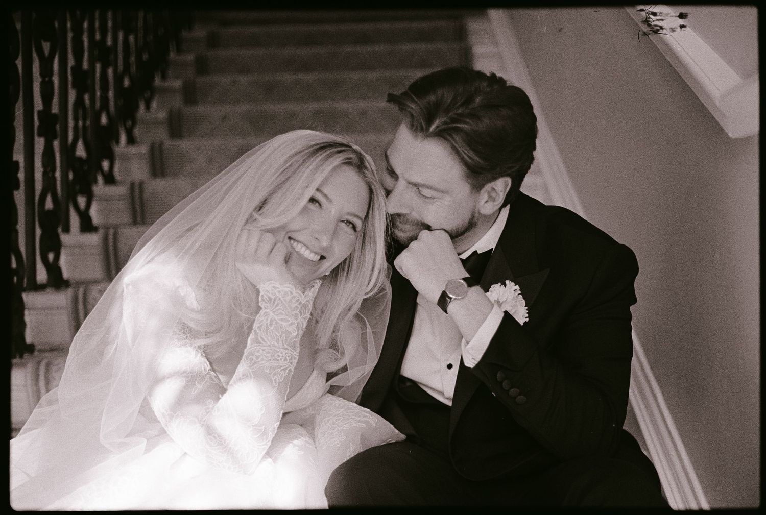 Black and white wedding photo of a smiling bride and groom sitting together on staircase steps.