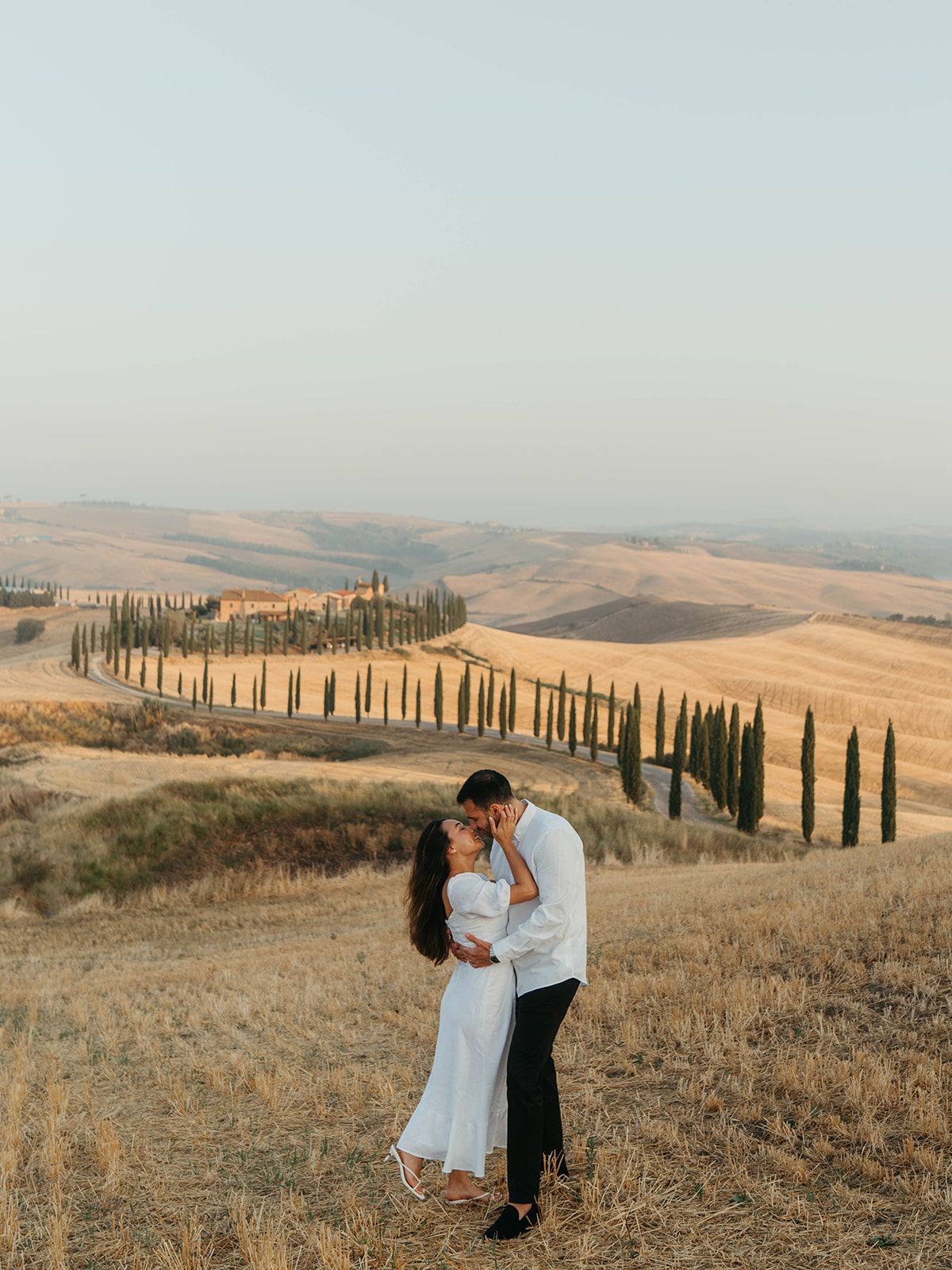 Couple sharing a romantic kiss in golden Tuscan hills with cypress tree-lined path and rolling countryside at sunset.