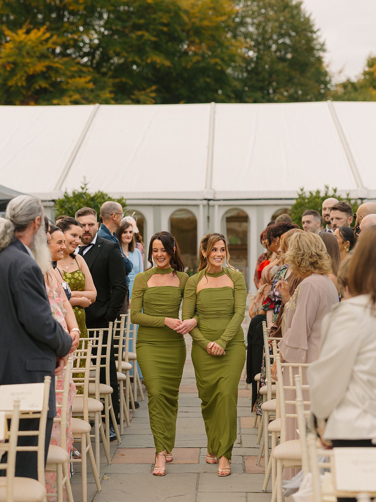 Wedding guests walk down the aisle in matching olive green dresses during an outdoor tent reception.