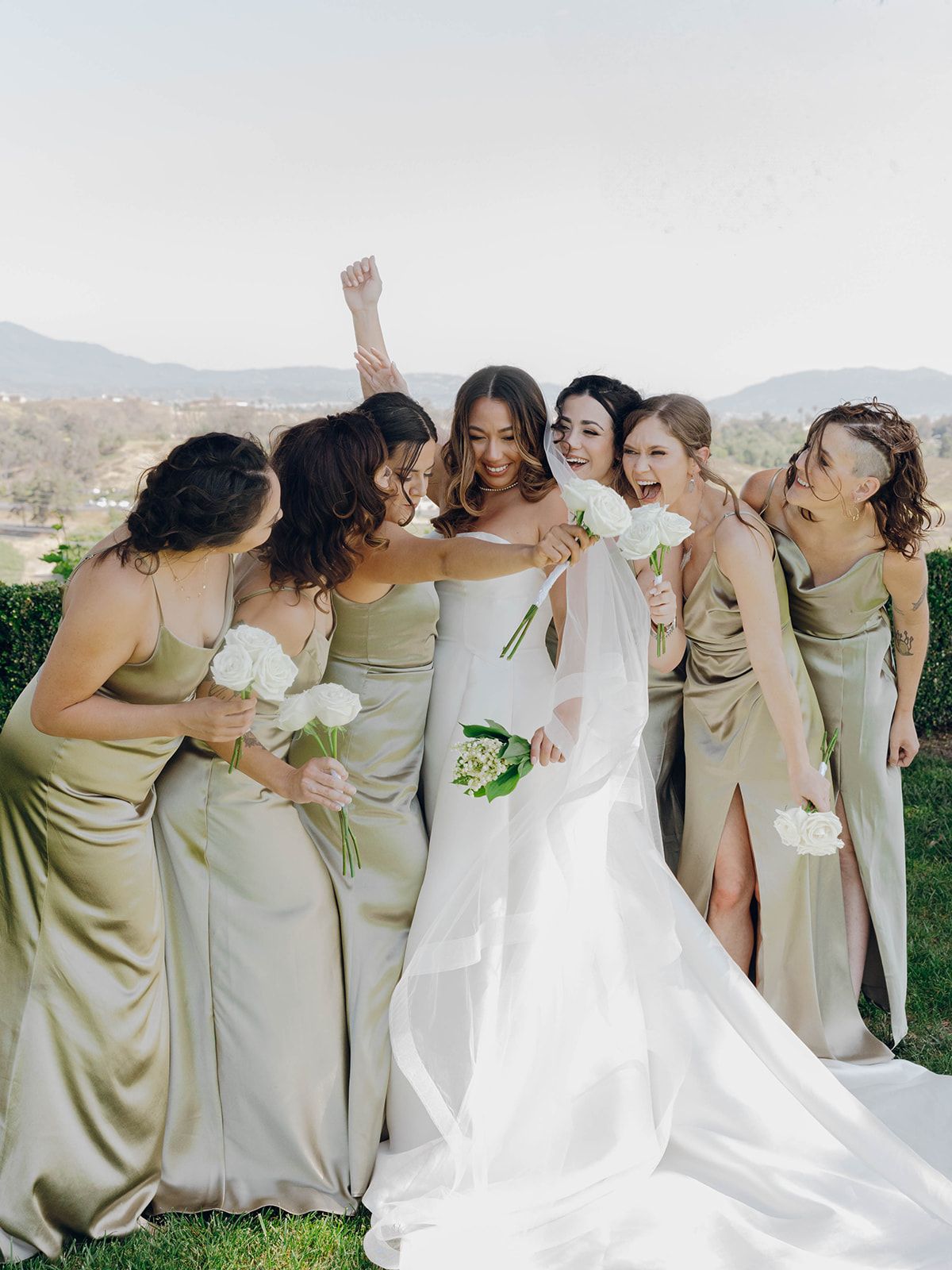 Bridesmaids in sage green silk dresses share a joyful moment with bride in white gown against mountain backdrop.