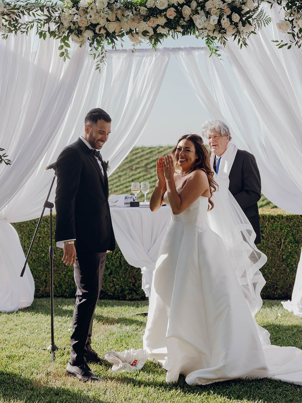 An emotional wedding ceremony takes place under a floral arch with white draping and vineyard views in the background.