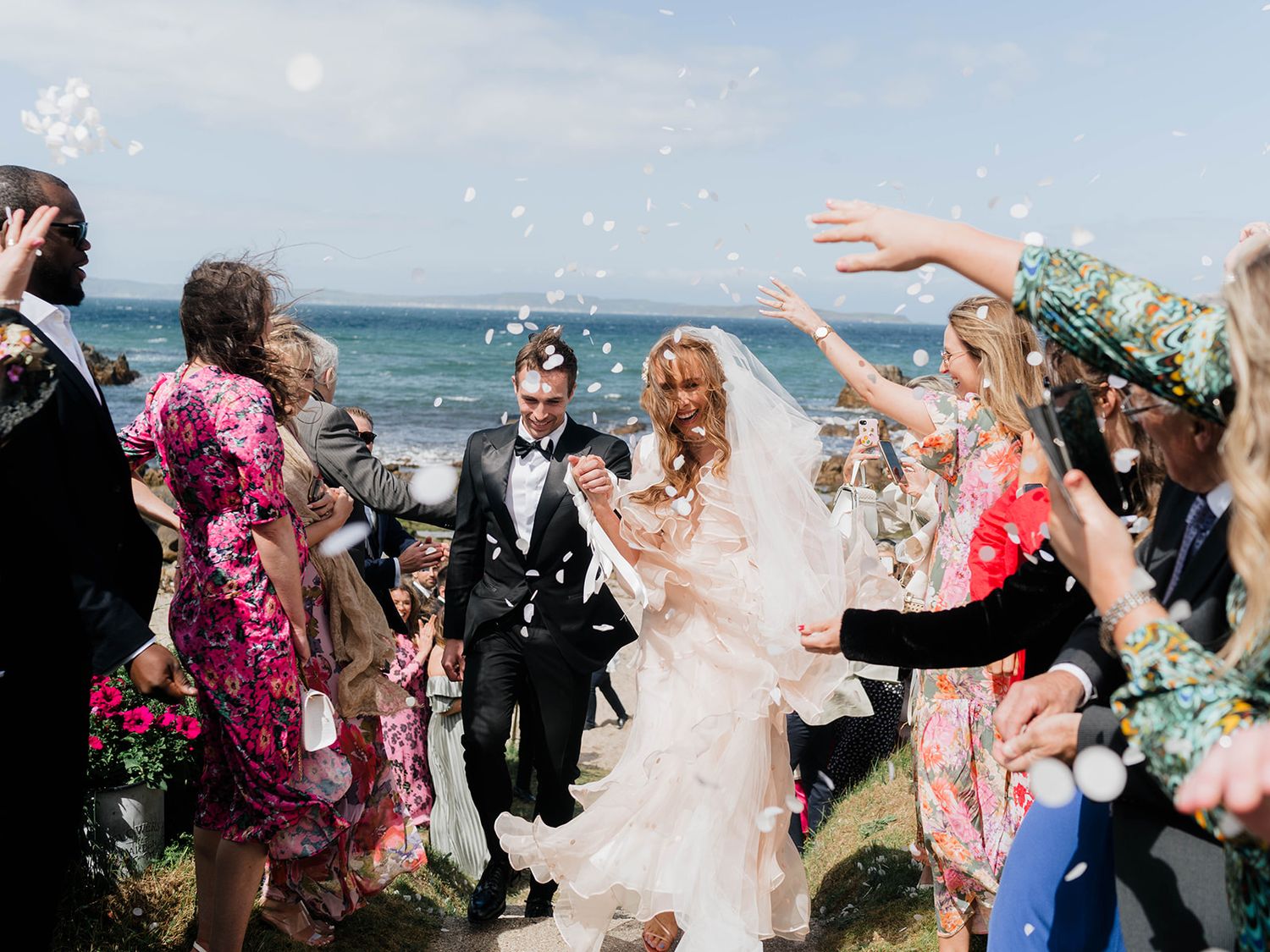 Wedding celebration on coastal cliffs with guests throwing confetti and bubbles.