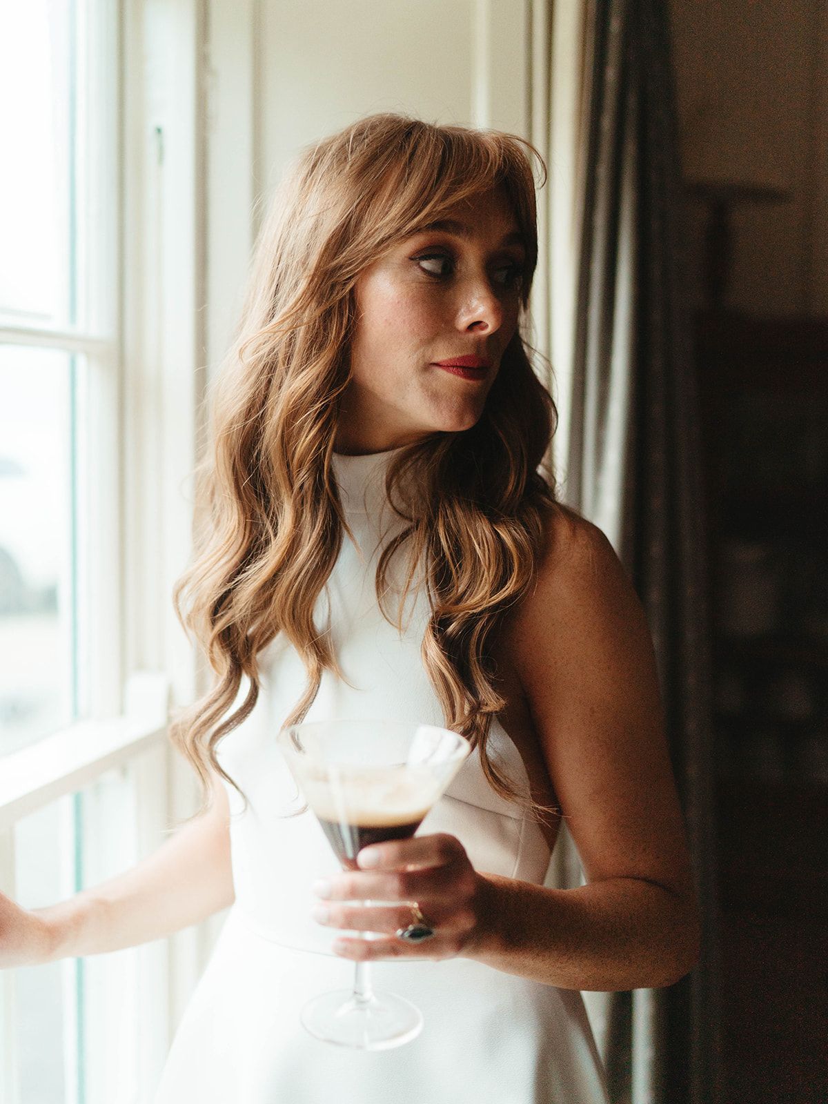 Bride in elegant white dress holds wine glass while gazing out window.