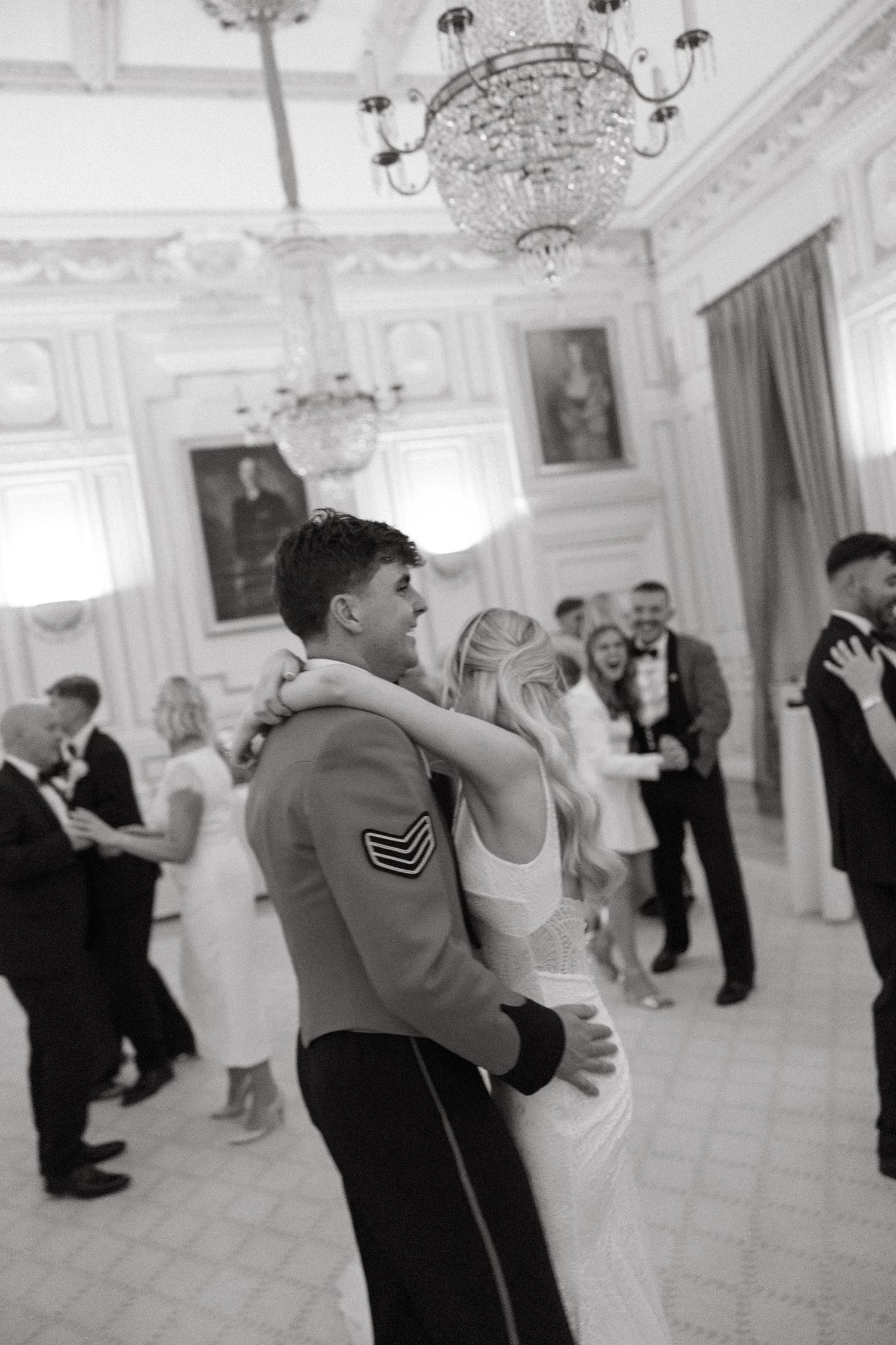 Couples dance elegantly in a grand ballroom with crystal chandeliers and ornate white decor during a formal celebration.