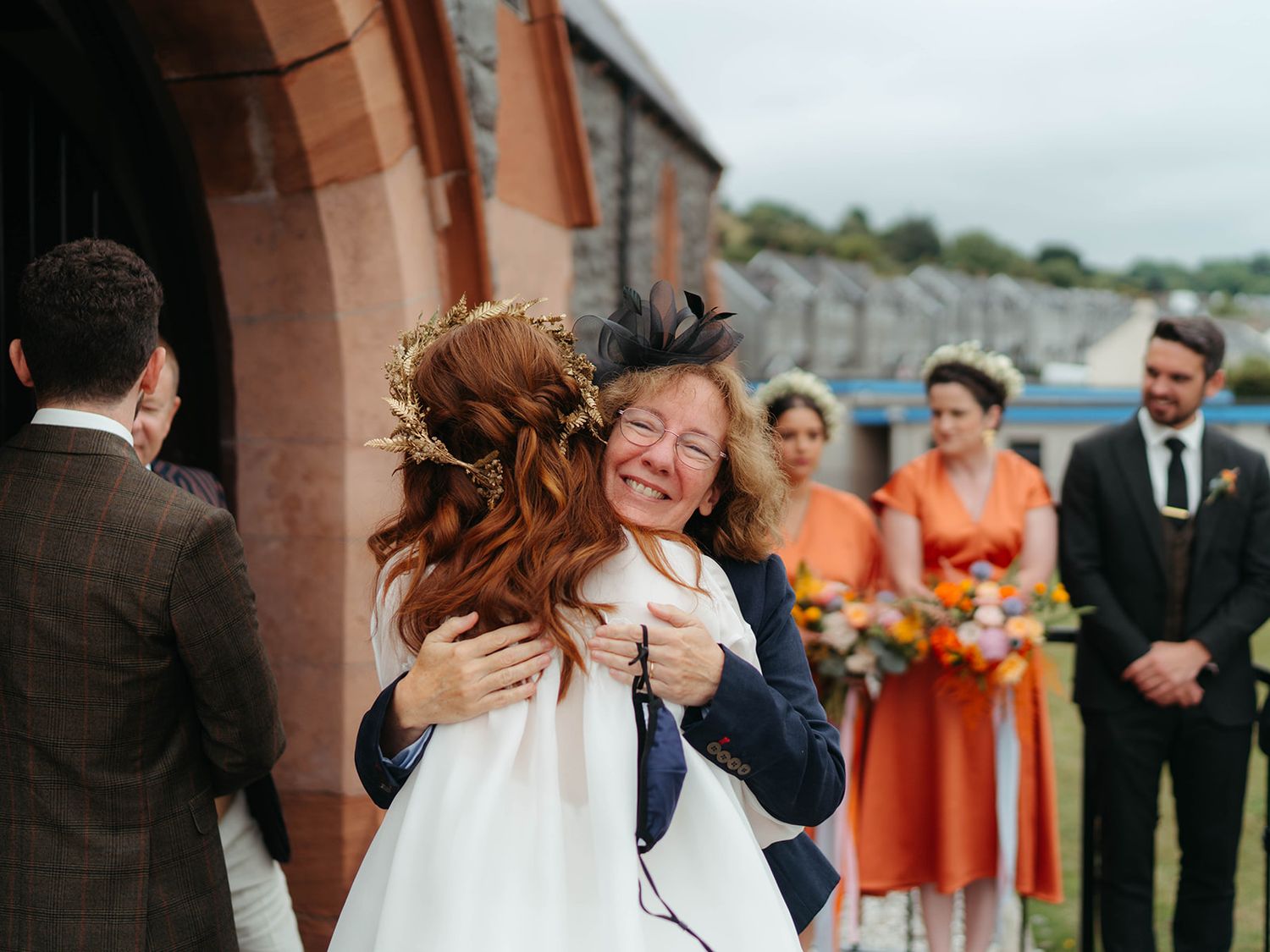 A warm embrace between two people at an outdoor wedding with bridesmaids in coral dresses looking on in the background.