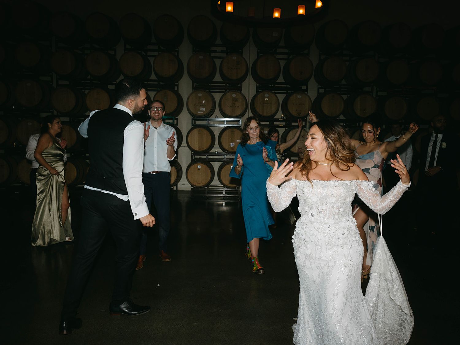 People dancing and celebrating at a wedding reception in front of wine barrels in a rustic cellar venue.