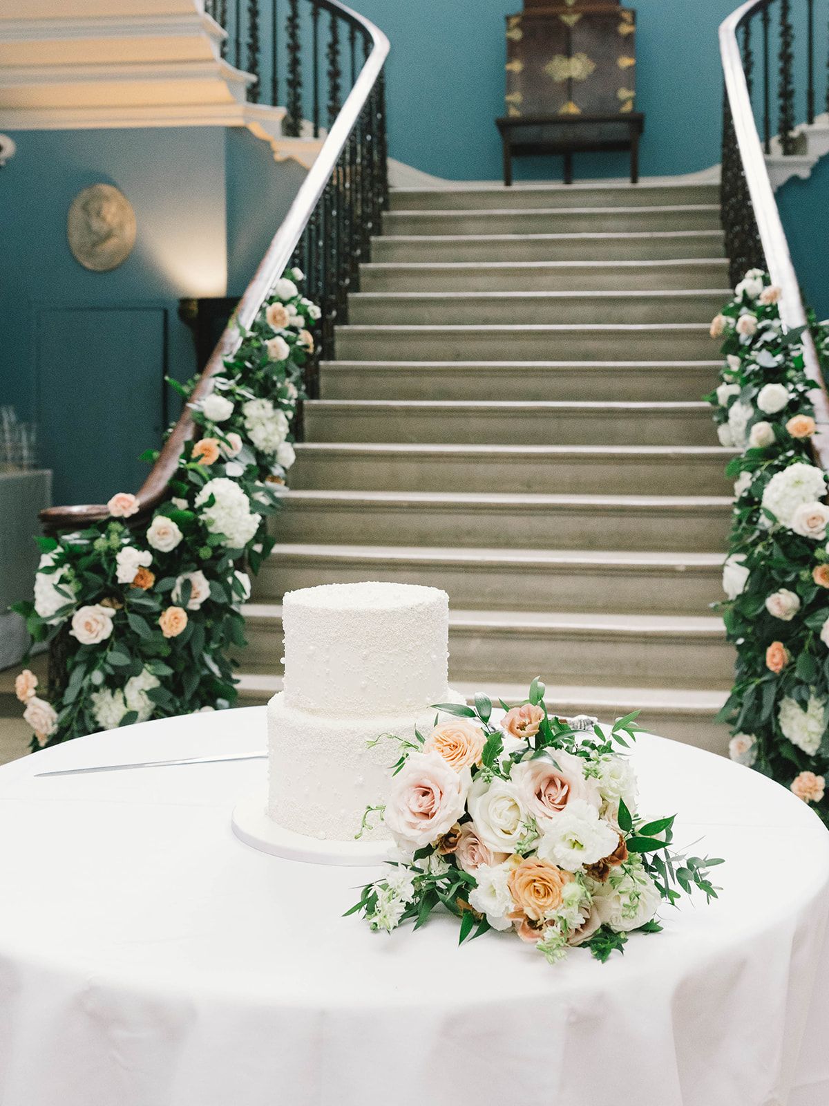 Elegant white wedding cake adorned with peach roses sits on white table near grand staircase decorated with floral garlands.