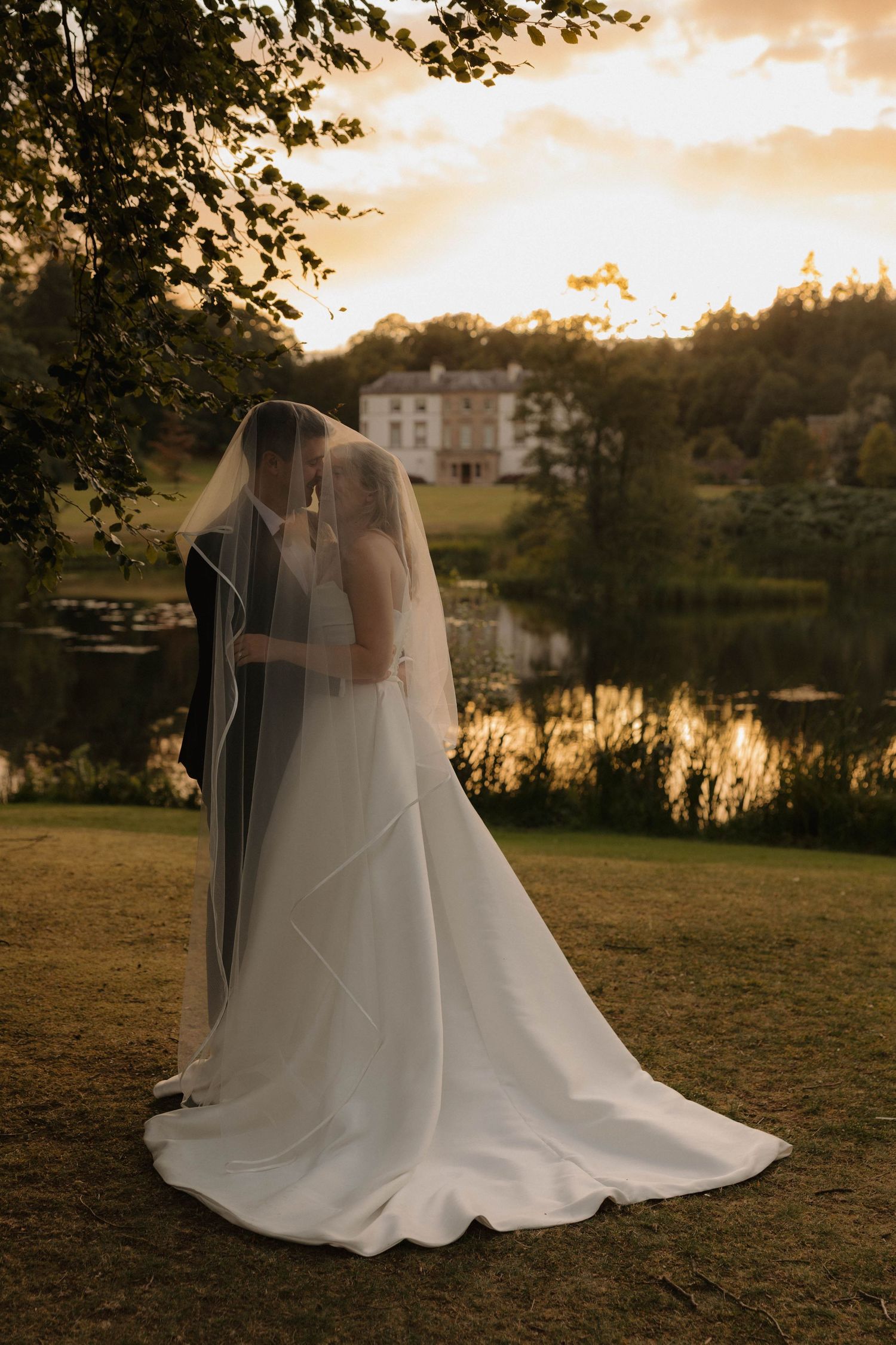 A wedding couple embraces at sunset by a lake with a grand estate house visible in the background.