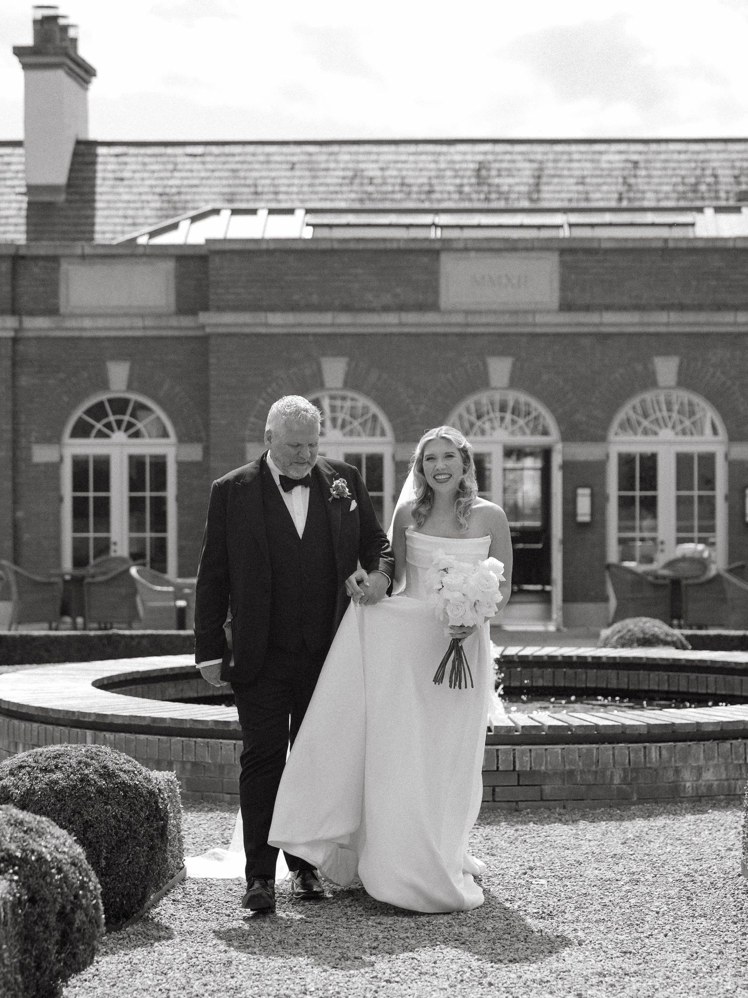 Wedding couple walks along a formal courtyard with brick buildings and fountain in background.