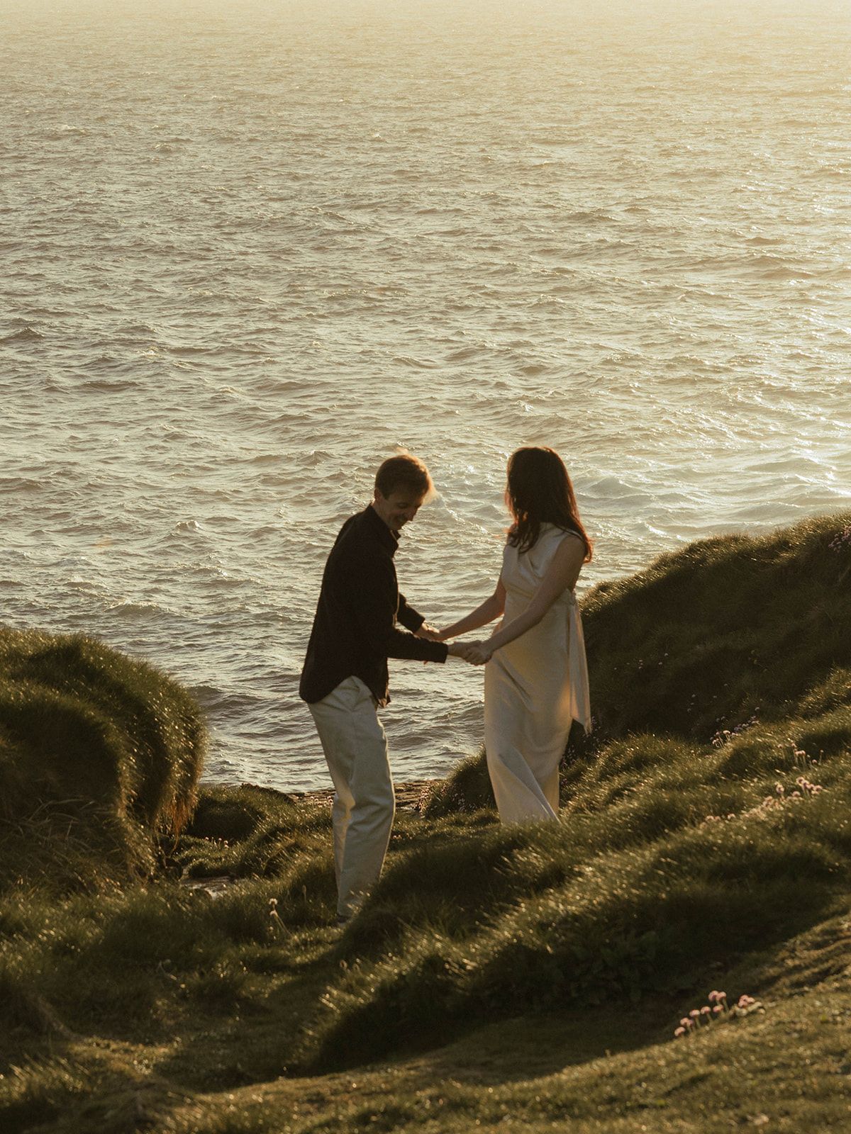 A couple walks along coastal cliffs during a golden sunset.