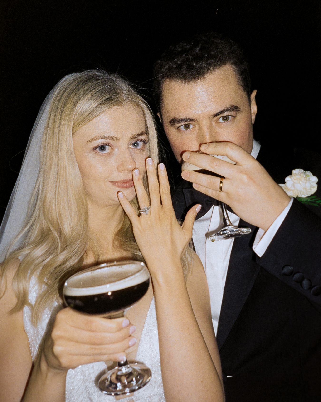 A bride and groom in formal wedding attire showing their rings and holding wine glasses while posing together.
