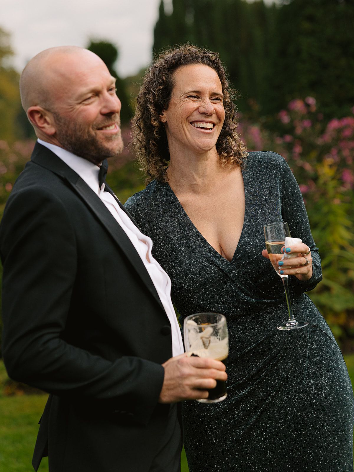 Couple in formal attire share a joyful moment while holding drinks in a garden setting.