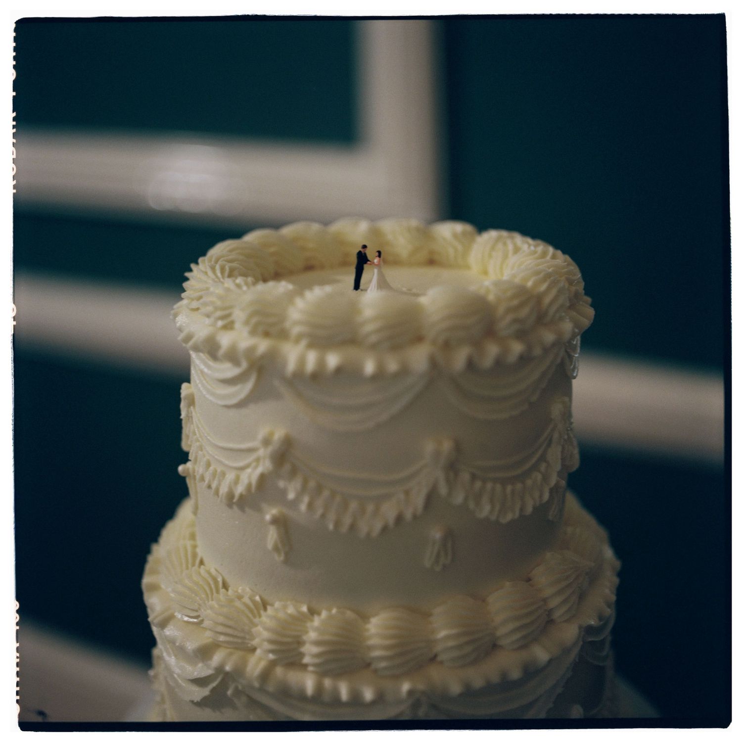 A two-tier white wedding cake with intricate frosting details and a bride and groom topper on top.